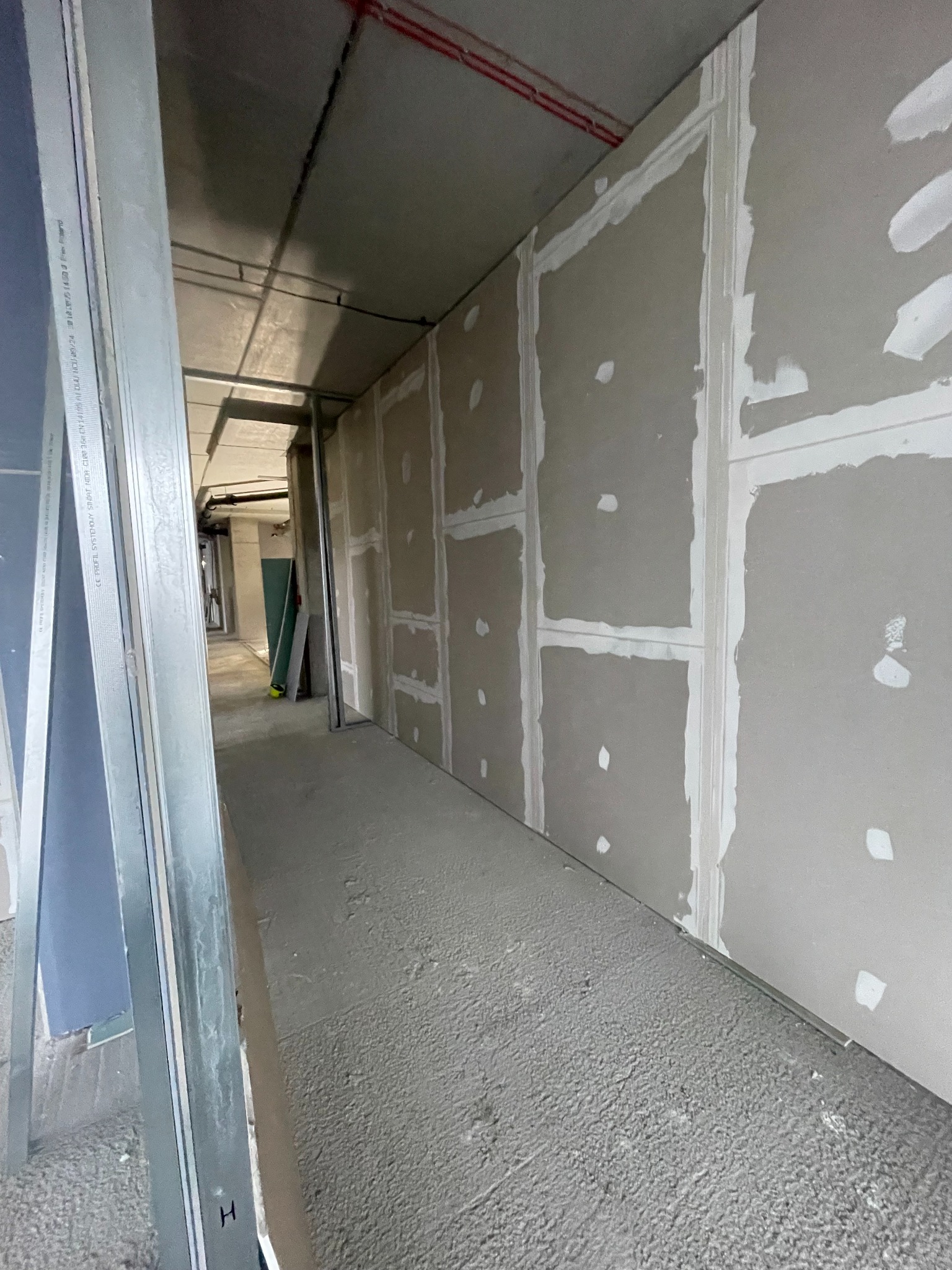 Korytarz w trakcie montażu płyt gipsowo-kartonowych. Widoczne metalowe profile i wstępnie wyszpachlowane łączenia płyt. Surowy beton na podłodze.