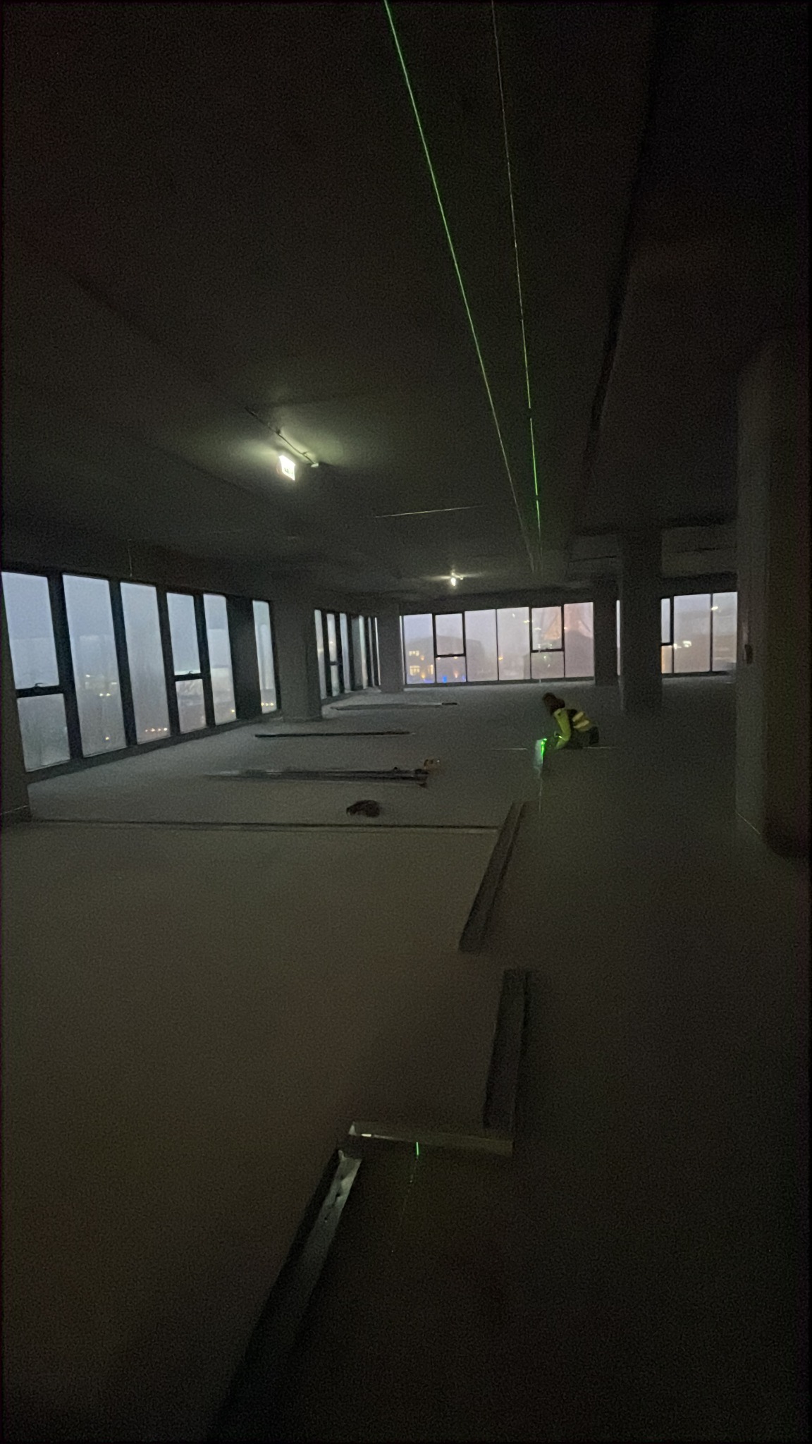 Wnętrze w trakcie prac wykończeniowych. Widoczne profile, poziomica laserowa wyznaczająca płaszczyzny. W tle okna z widokiem na miasto i pracownik w kamizelce.