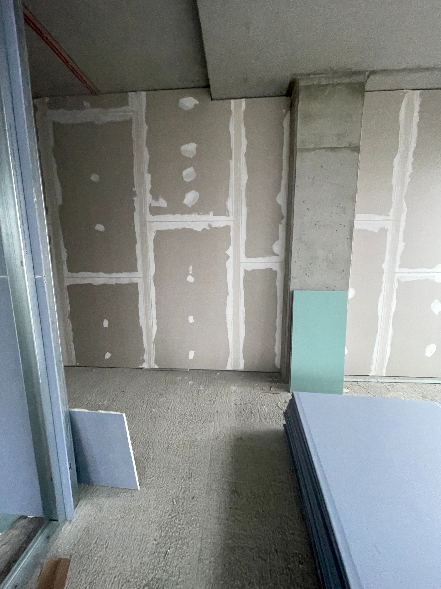 Montaż płyt gipsowo-kartonowych na ścianie z widocznymi łączeniami i szpachlowaniem. Na podłodze stos płyt. Widoczny betonowy filar.