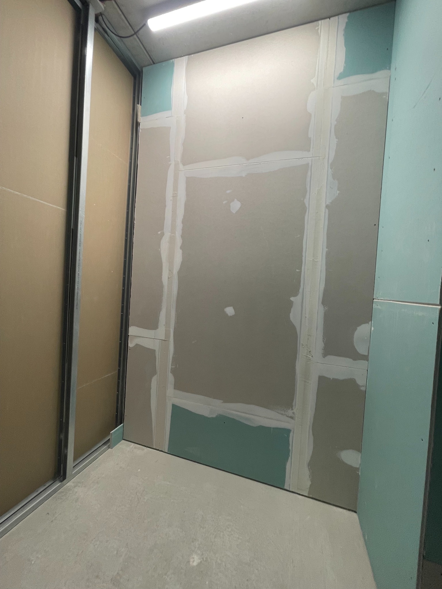 Ściana z płyt gipsowo-kartonowych w trakcie montażu, widoczne łączenia szpachlowane na biało, fragmenty zielonych płyt. Metalowa konstrukcja w tle.