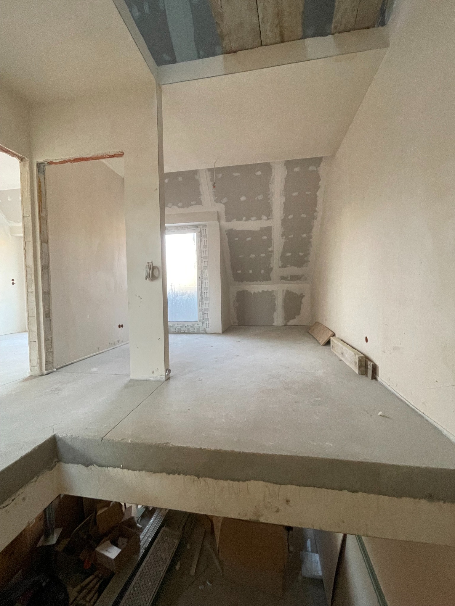 Surowe wnętrze w trakcie prac wykończeniowych: ściany z płyt gipsowo-kartonowych, surowa podłoga, widoczne elementy konstrukcyjne i okno z widokiem na zewnątrz.