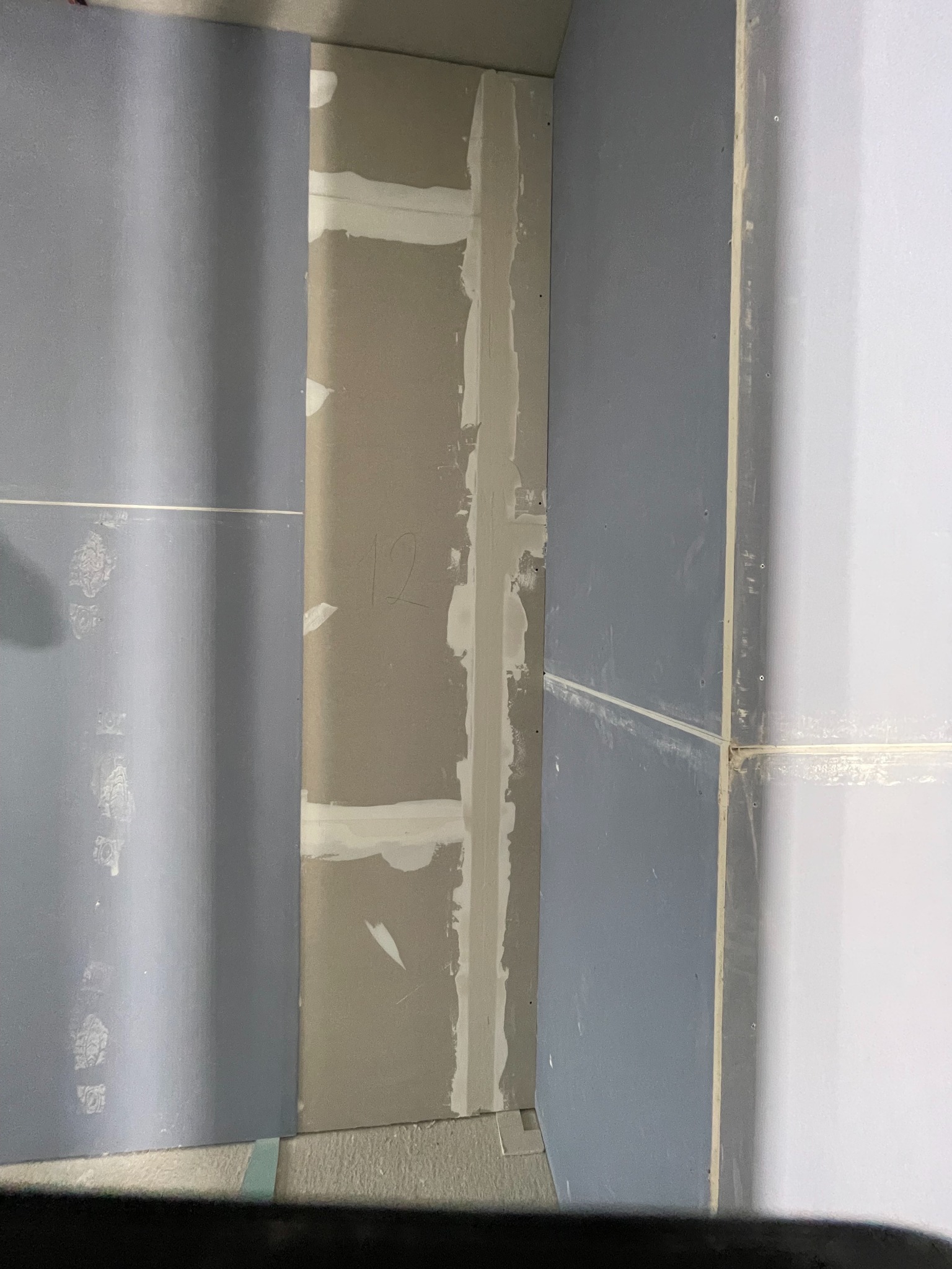 Fragment ściany z płyt kartonowo-gipsowych, widoczne łączenia płyt wypełnione masą szpachlową. Na jednej z płyt widnieje napis '12'.