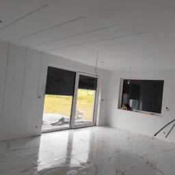 Marcel Woźniak - Jasne wnętrze w trakcie wykańczania: białe ściany z listwami, marmurowe płytki podłogowe, duże okno przesuwne z roletami. Widoczne przewody i niedokończone detale.