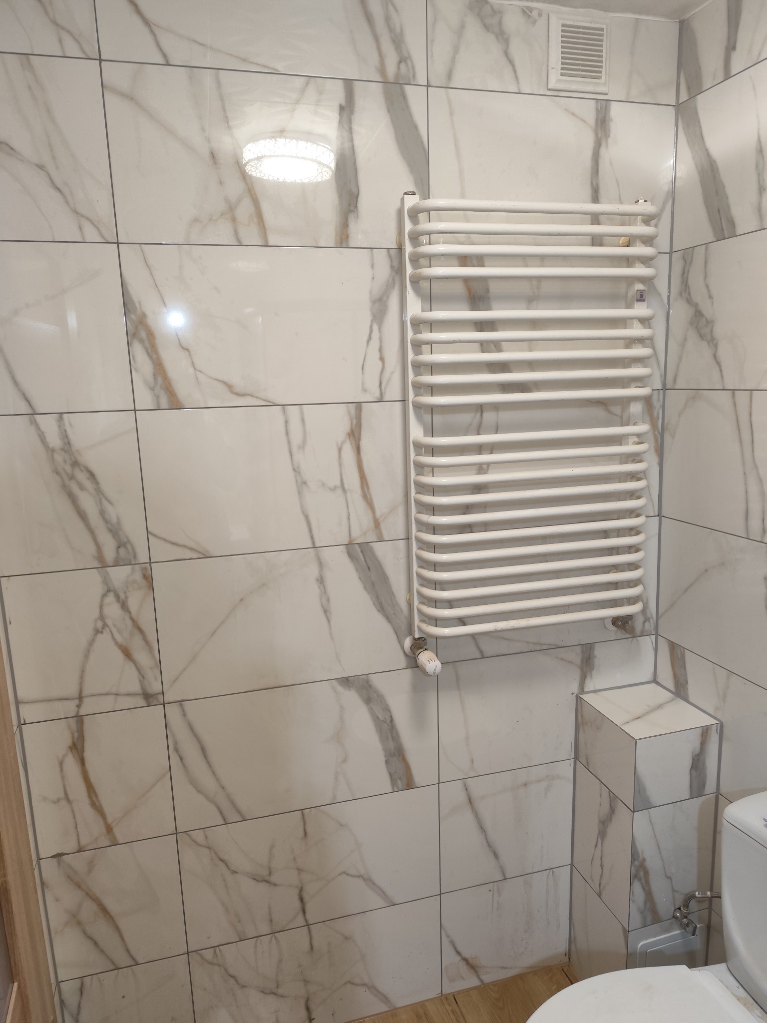 Wykończona łazienka z białymi płytkami imitującymi marmur i grzejnikiem drabinkowym. Widoczny fragment toalety i wentylacja. Ujęcie z dołu, podkreślające detale.