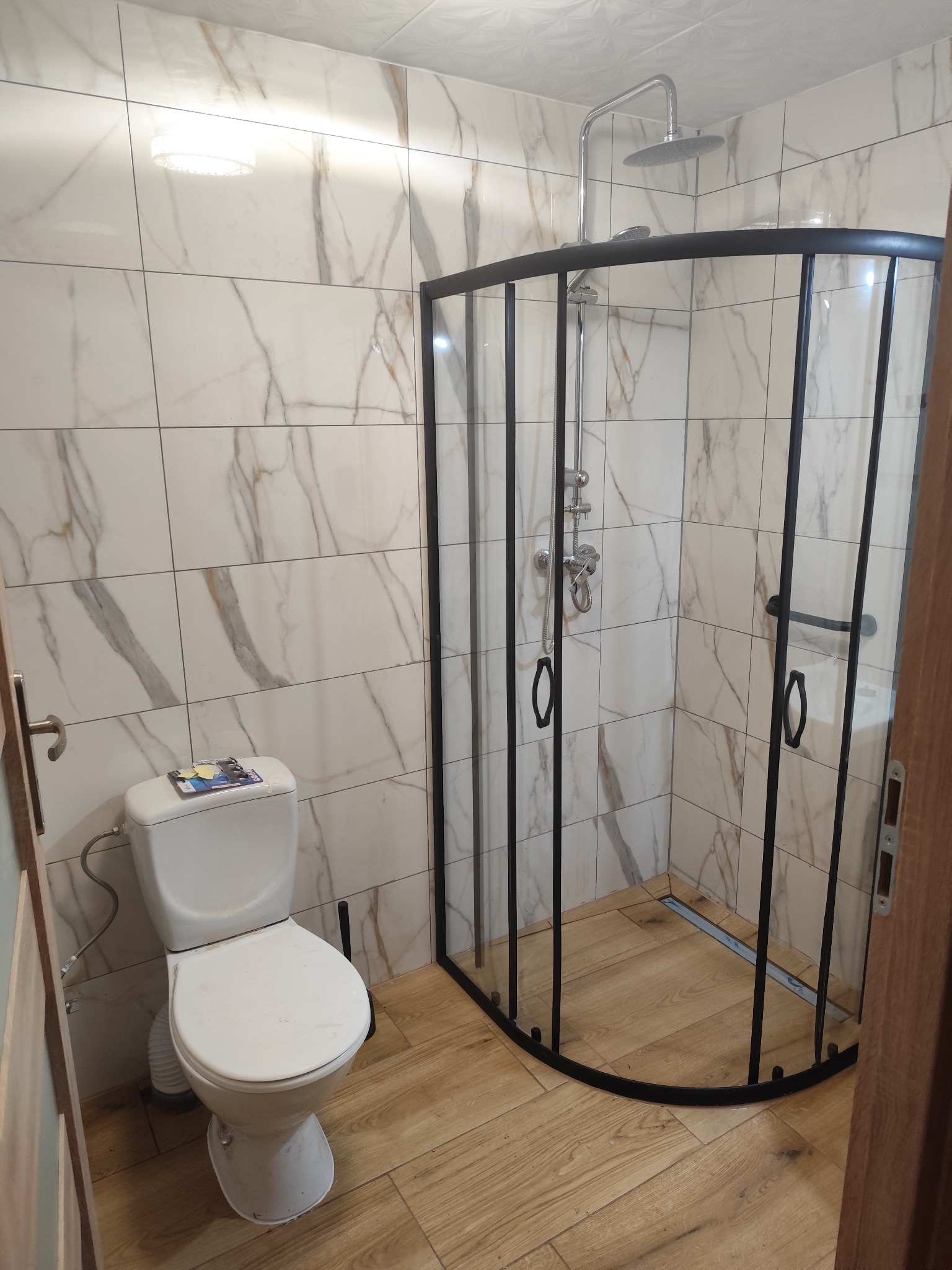 Wykończona łazienka z kabiną prysznicową typu walk-in z czarnymi profilami, toaletą i płytkami imitującymi marmur. Jasne drewnopodobne panele na podłodze.