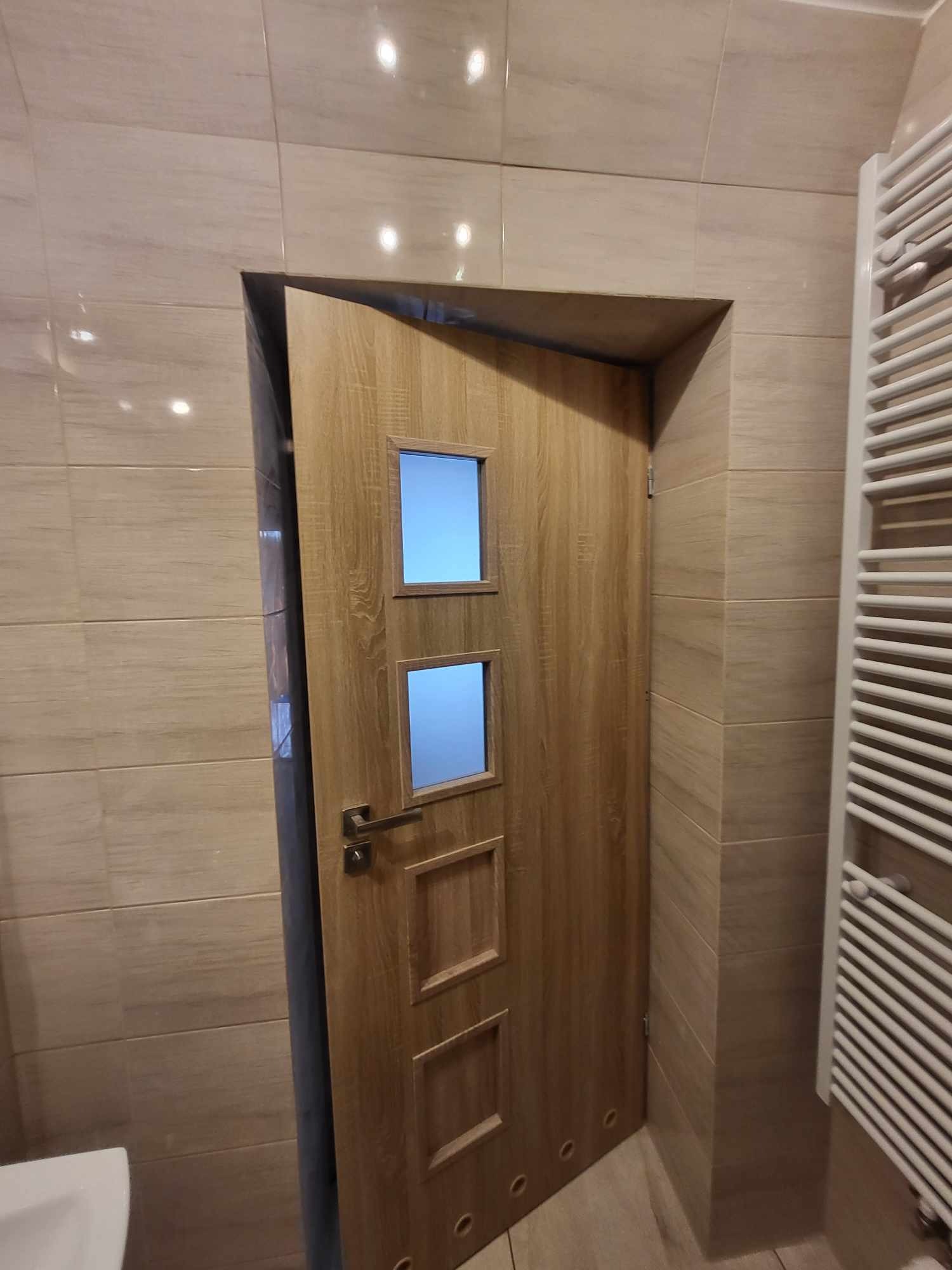 Otwarta, drewniana, wewnętrzna  ościeżnica z trzema przeszkleniami w łazience wyłożonej beżowymi płytkami. Widoczny grzejnik drabinkowy.