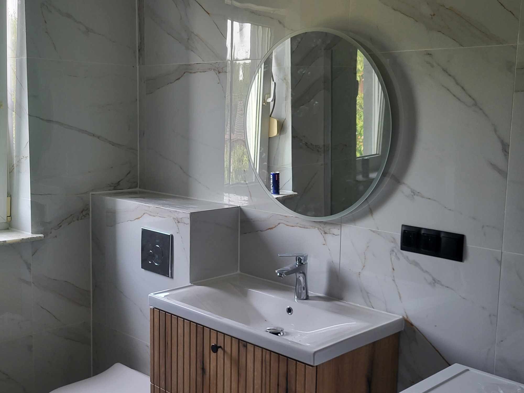 Wykończona łazienka z marmurowymi płytkami, okrągłym lustrem i drewnianą szafką pod umywalką. Widoczne detale: bateria, włączniki i przycisk spłuczki.