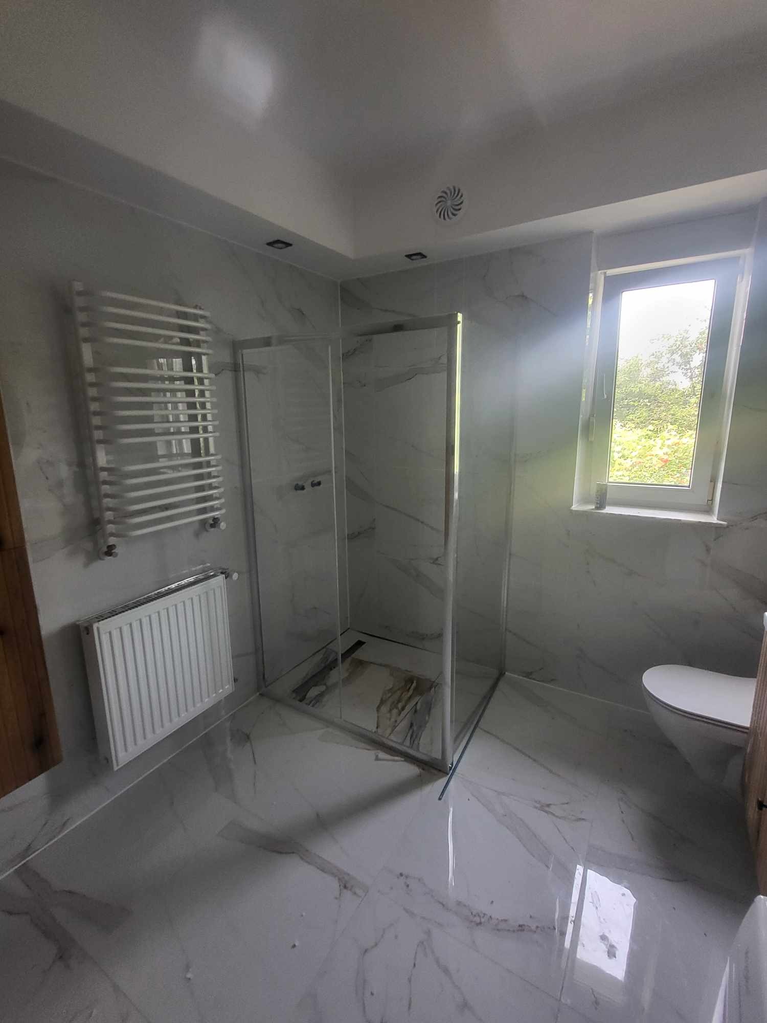 Wykończona łazienka w bieli: kabina prysznicowa ze srebrnymi ramami, grzejnik drabinkowy, marmurowe płytki i okno z widokiem na zieleń. Nowoczesny design.