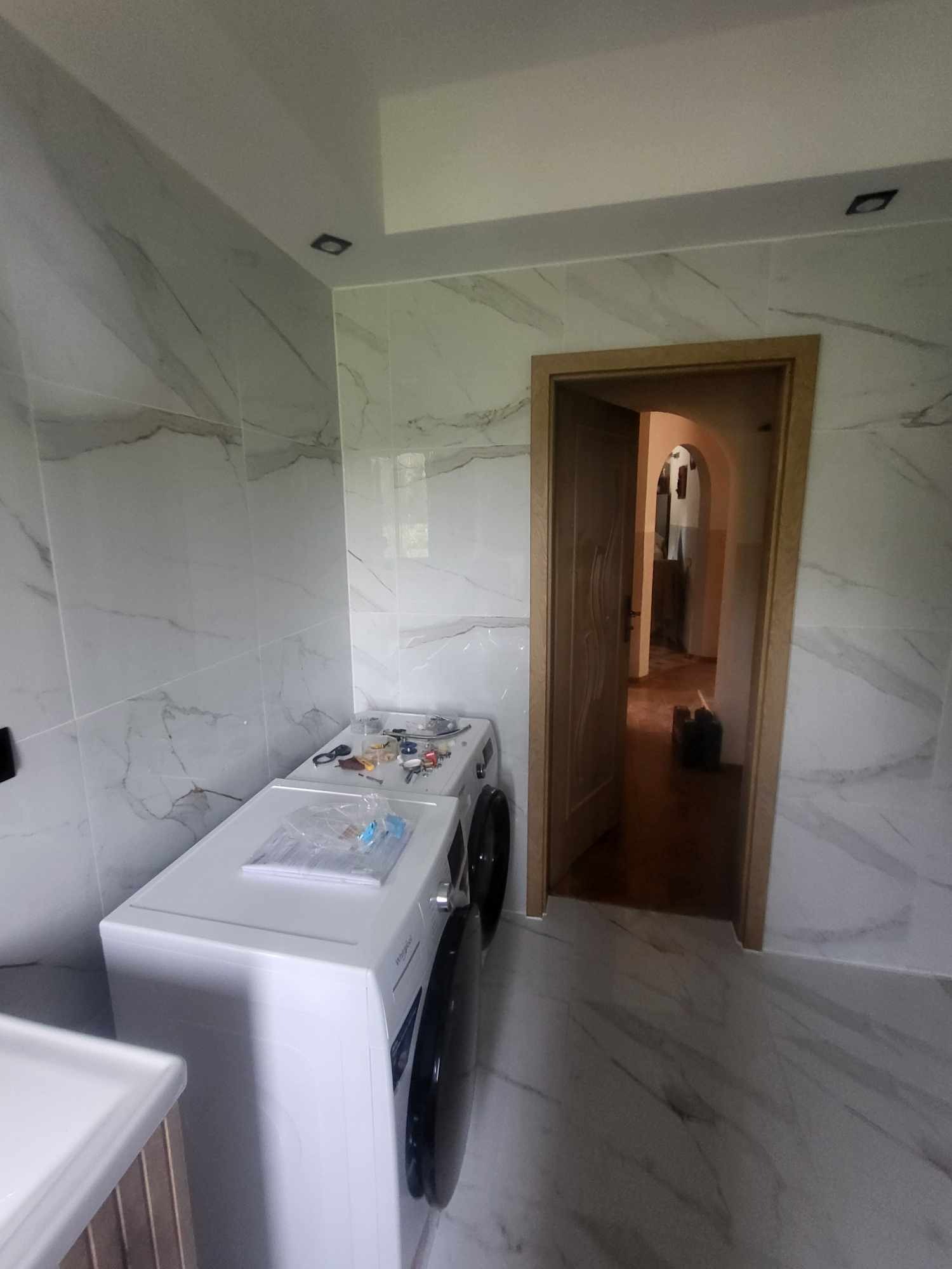 Wykończona łazienka z marmurowymi płytkami, pralką i otwartymi drzwiami. Widoczne narzędzia na pralce. Nowoczesny design, jasne kolory, świeżo po remoncie.