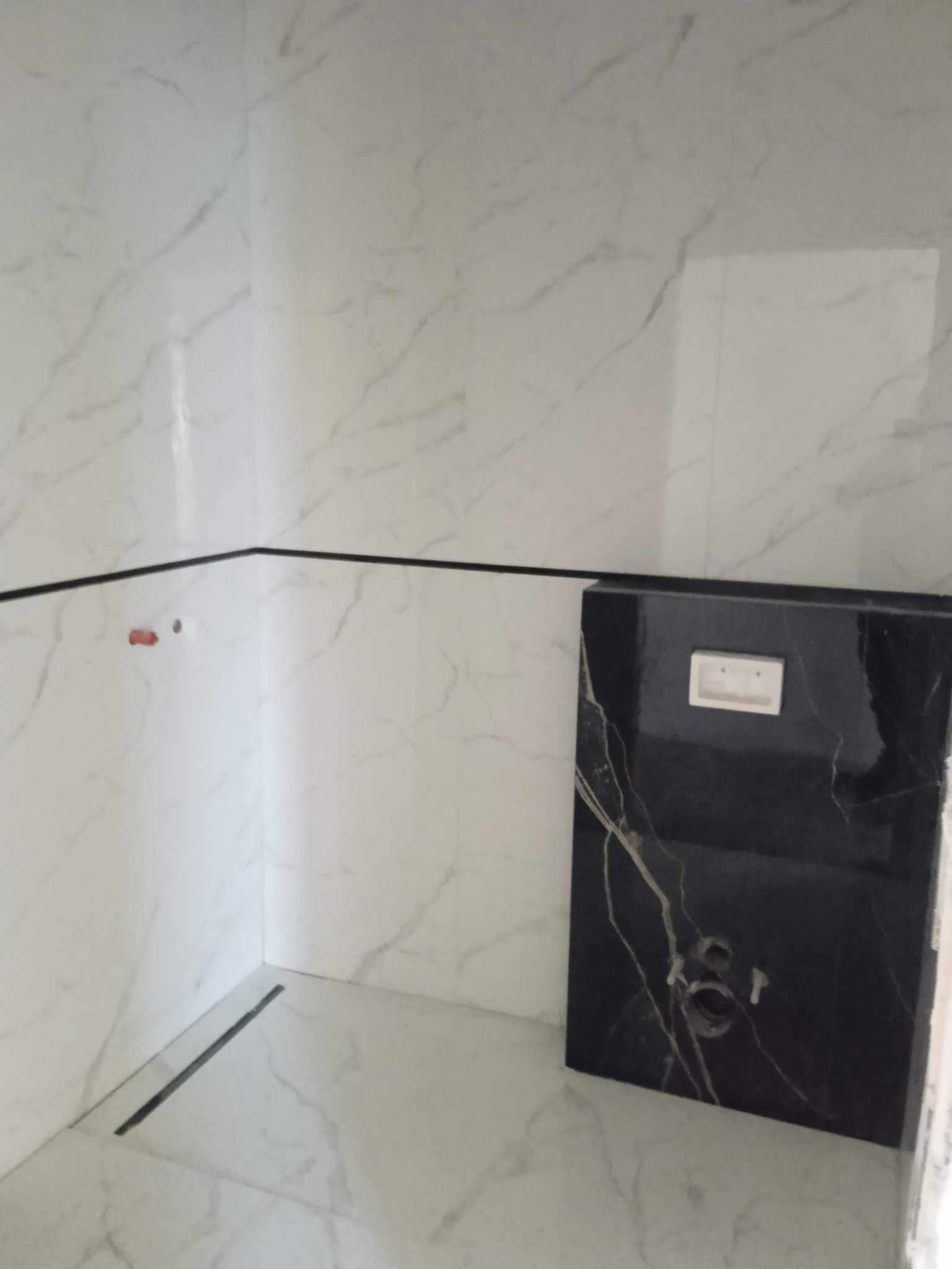 Wykończona łazienka z białymi płytkami imitującymi marmur i czarną zabudową WC. Widoczny odpływ liniowy w podłodze. Minimalistyczny design z akcentem na kontrast kolorów.