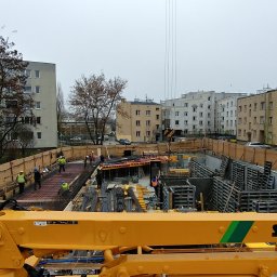 PAROTECH SPÓŁKA Z OGRANICZONĄ ODPOWIEDZIALNOŚCIĄ - Budowa fundamentów pod budynek wielorodzinny w otoczeniu bloków. Widoczna zbrojona konstrukcja, pracownicy w kaskach i żółty wysięgnik pompy do betonu Schwing S28+4.