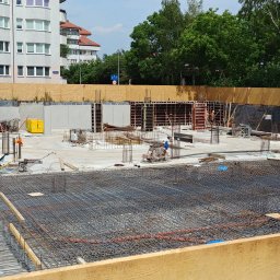 PAROTECH SPÓŁKA Z OGRANICZONĄ ODPOWIEDZIALNOŚCIĄ - Zbrojenie fundamentów pod budowę. Widoczne stalowe pręty, szalunki i betonowe ściany. W tle budynki mieszkalne i drzewa. Prace budowlane w toku.