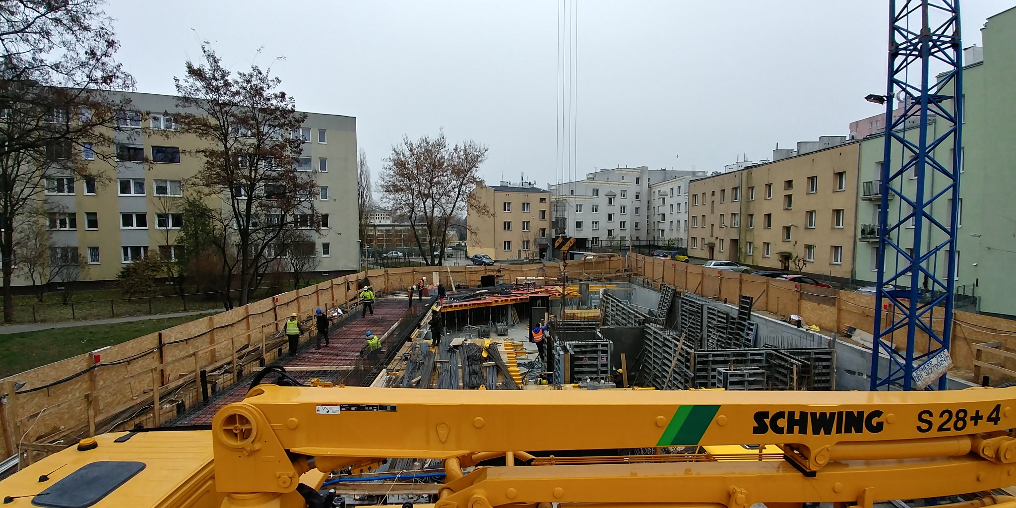 Budowa fundamentów pod budynek wielorodzinny w otoczeniu bloków. Widoczna zbrojona konstrukcja, pracownicy w kaskach i żółty wysięgnik pompy do betonu Schwing S28+4.