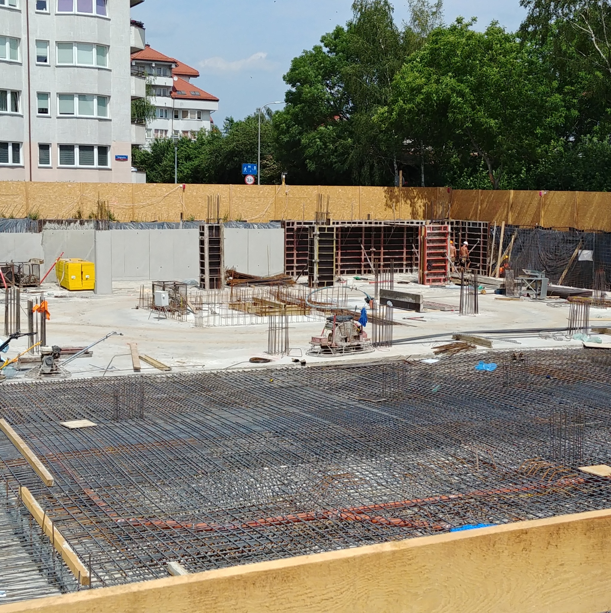 Zbrojenie fundamentów pod budowę. Widoczne stalowe pręty, szalunki i betonowe ściany. W tle budynki mieszkalne i drzewa. Prace budowlane w toku.
