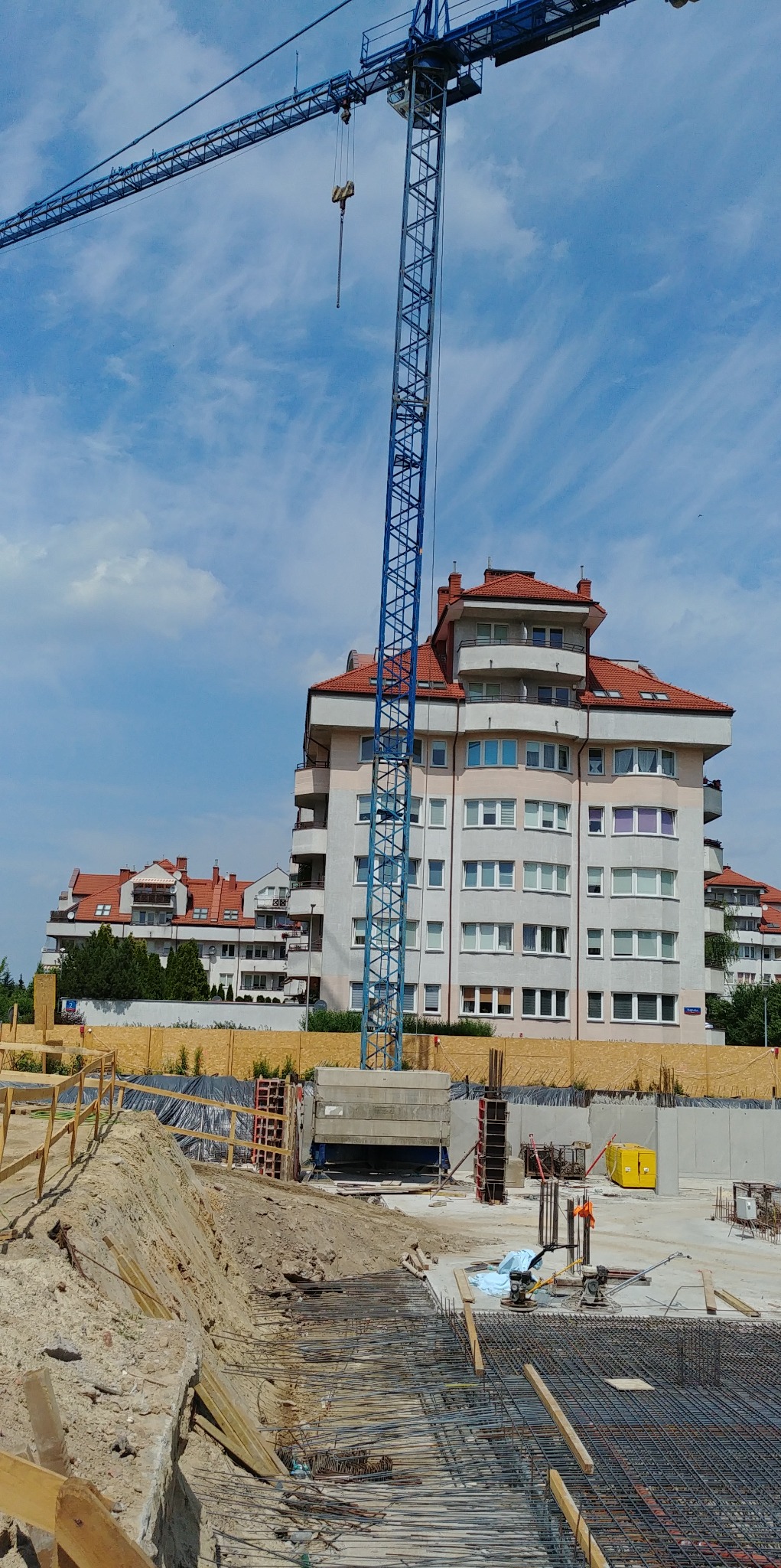 Budowa wielopiętrowego budynku mieszkalnego z czerwonym dachem. Na pierwszym planie widoczny żuraw budowlany i zbrojenie fundamentów. Słoneczny dzień.