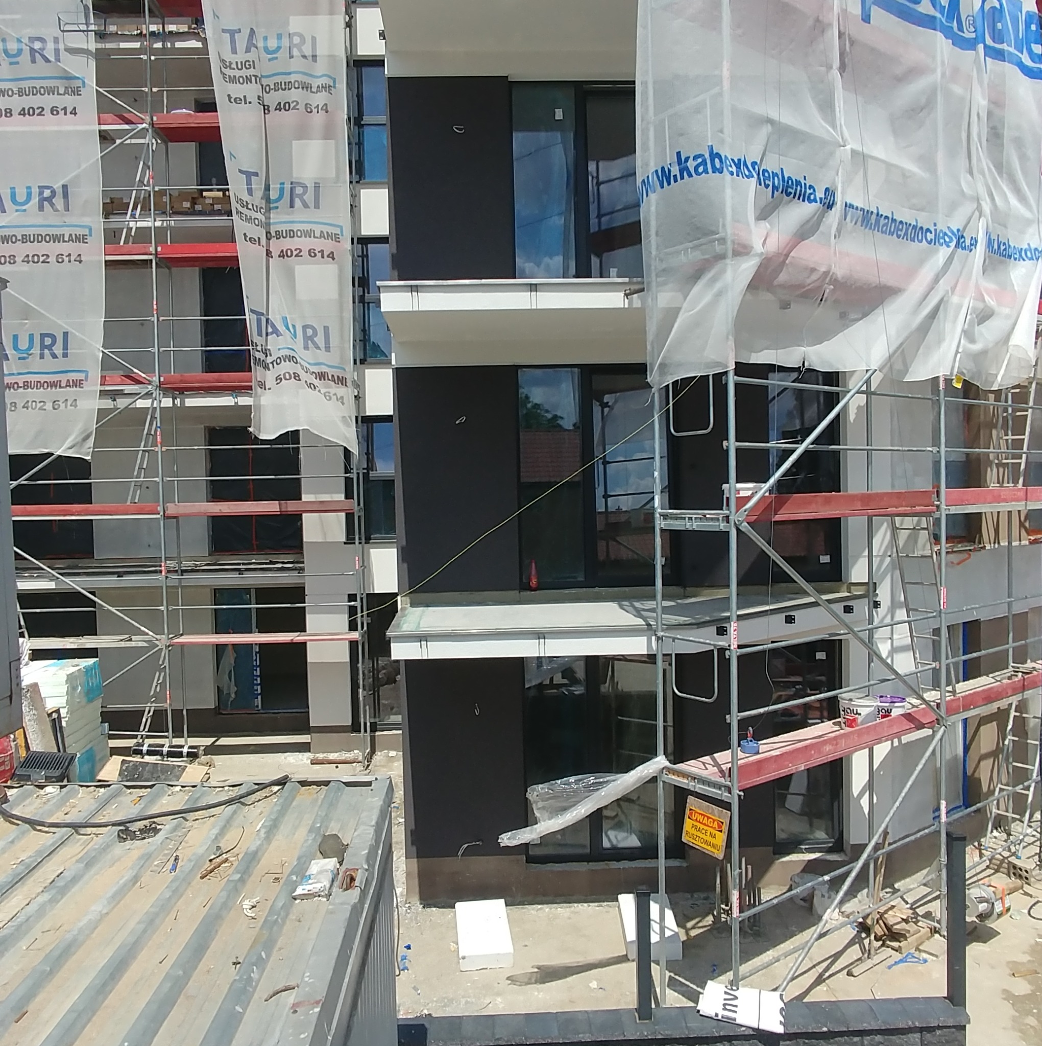 Wielopiętrowy budynek w trakcie prac wykończeniowych, z widocznymi rusztowaniami i materiałami budowlanymi. Częściowo zasłonięty banerami reklamowymi.