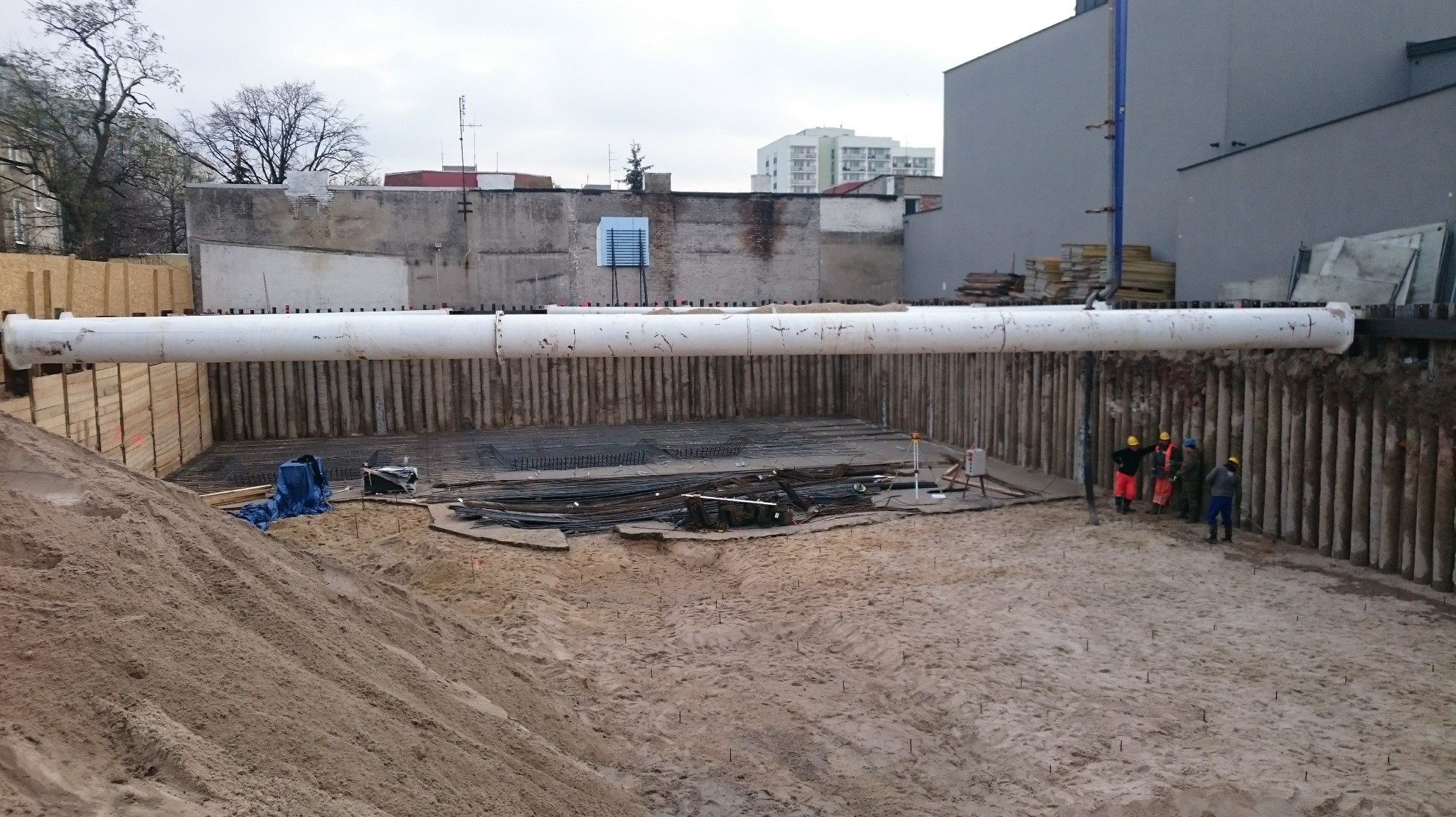 Wykonanie wykopu pod fundamenty z zabezpieczeniem ścianek palisadą. Widoczne zbrojenie i pracownicy w kaskach. Palisada wzmocniona rurą stalową.