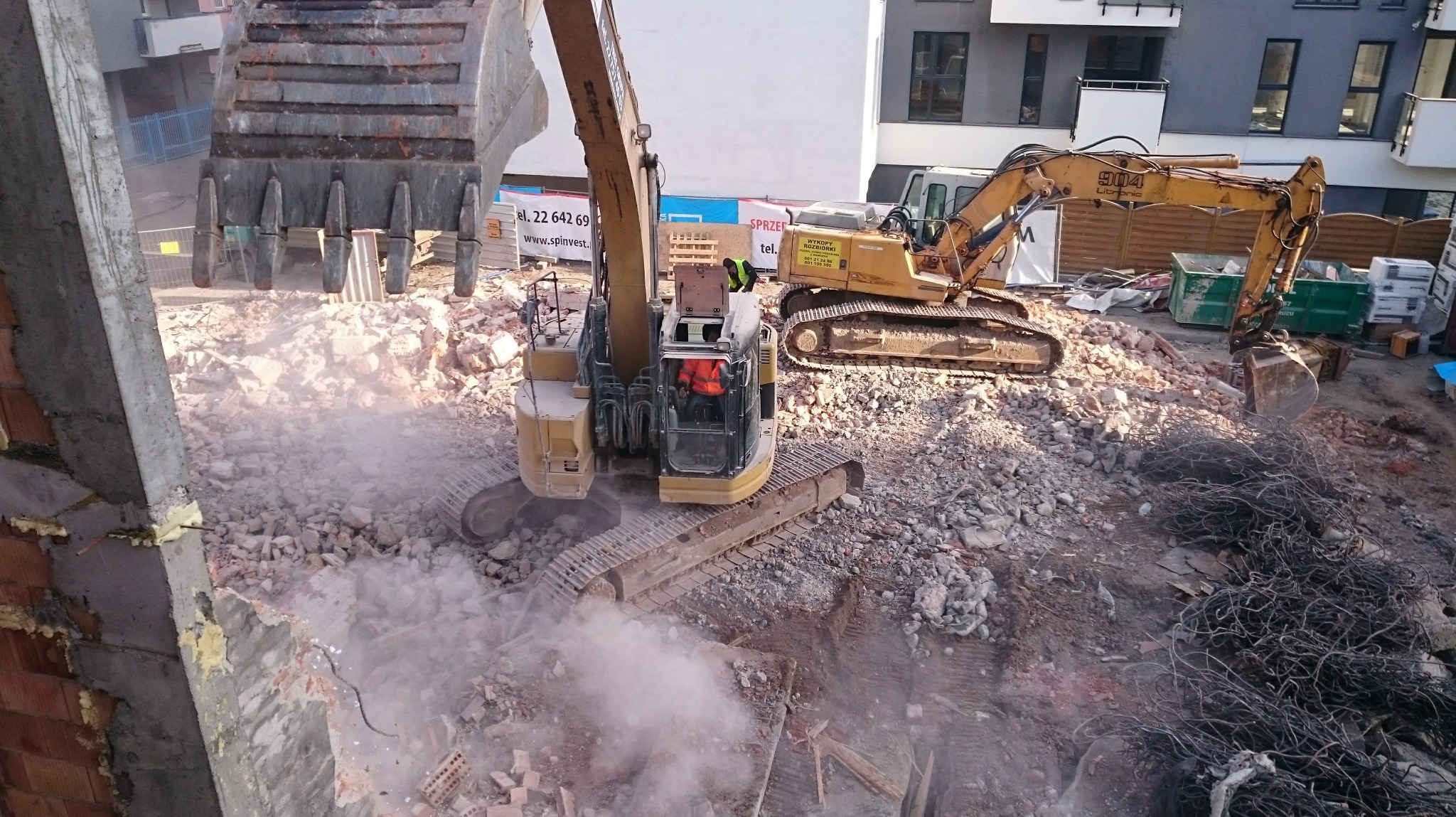 Dwa żółte, gąsienicowe koparki na placu budowy, gruz i zbrojenia, w tle budynek mieszkalny. Jedna koparka w trakcie pracy, unosi gruz. Widok z góry.