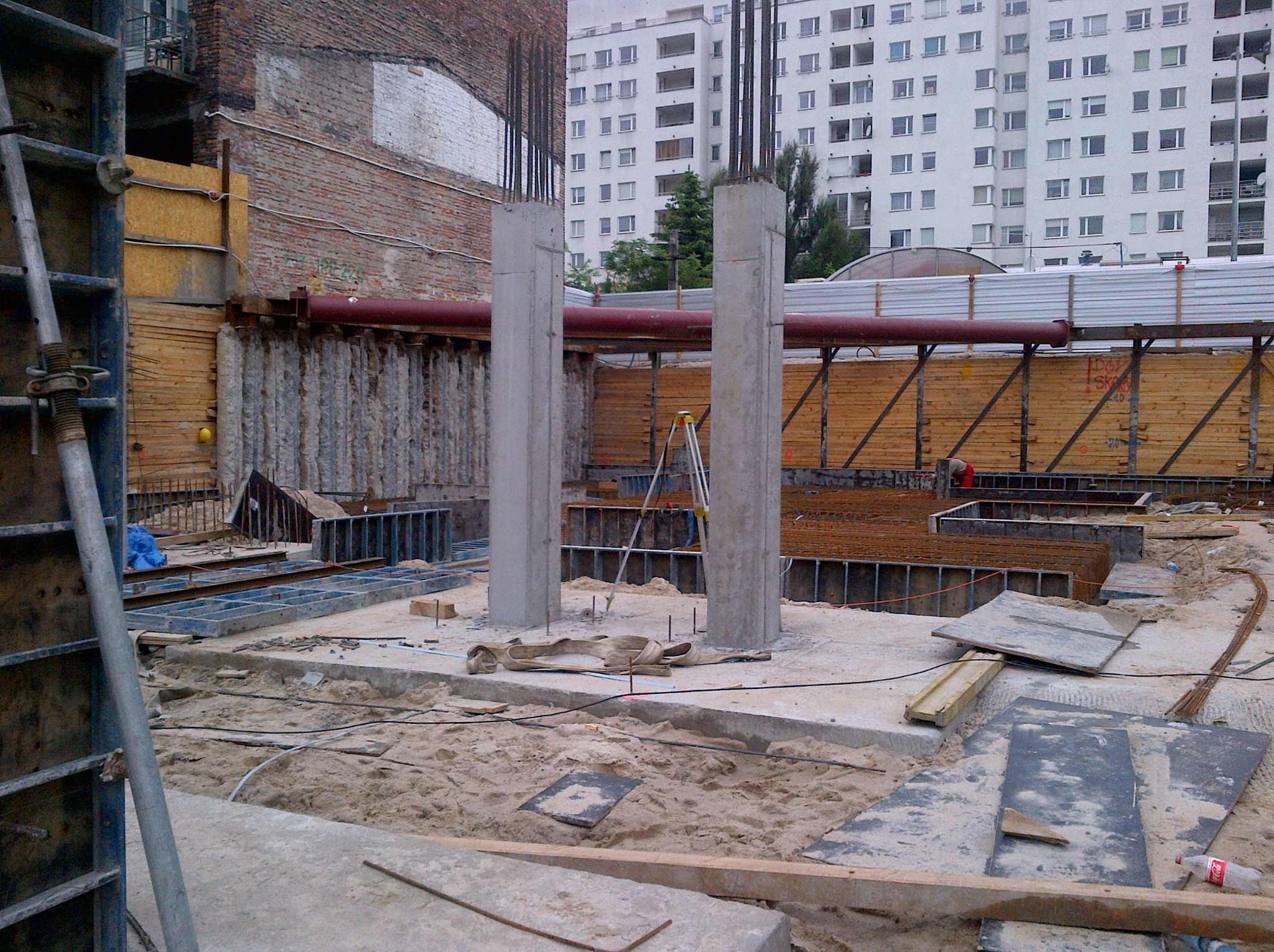 Wykop fundamentów z widocznymi szalunkami, zbrojeniami i dwoma betonowymi słupami na tle budynku mieszkalnego i ceglanej ściany. Widoczne zabezpieczenia wykopu.