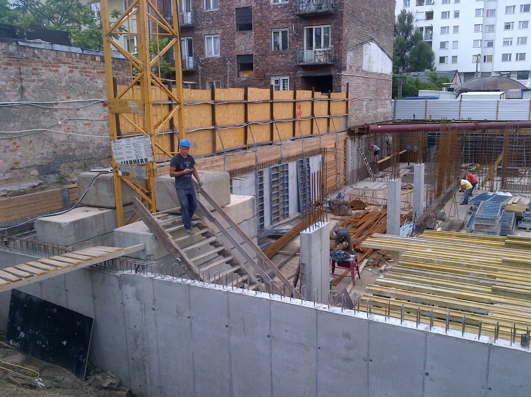 Budowa fundamentów z betonu i zbrojenia na placu budowy. Widoczny pracownik w niebieskim kasku i dźwig Liebherr. W tle budynki mieszkalne.