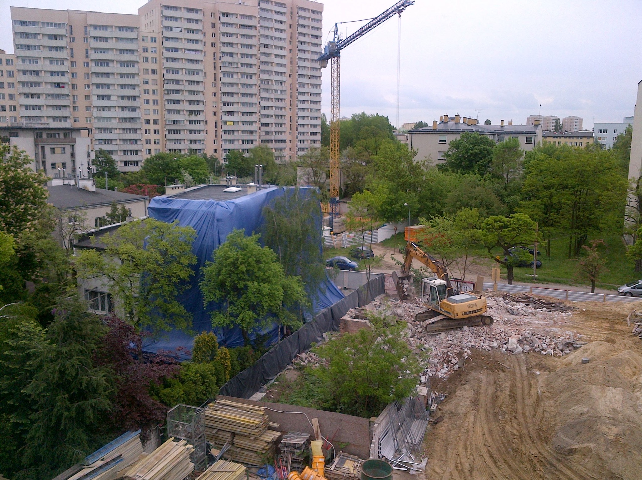 Prace rozbiórkowe z użyciem koparki na tle budynku mieszkalnego i żurawia budowlanego. Część budynku zabezpieczona niebieską plandeką. Widoczne składowane materiały budowlane.