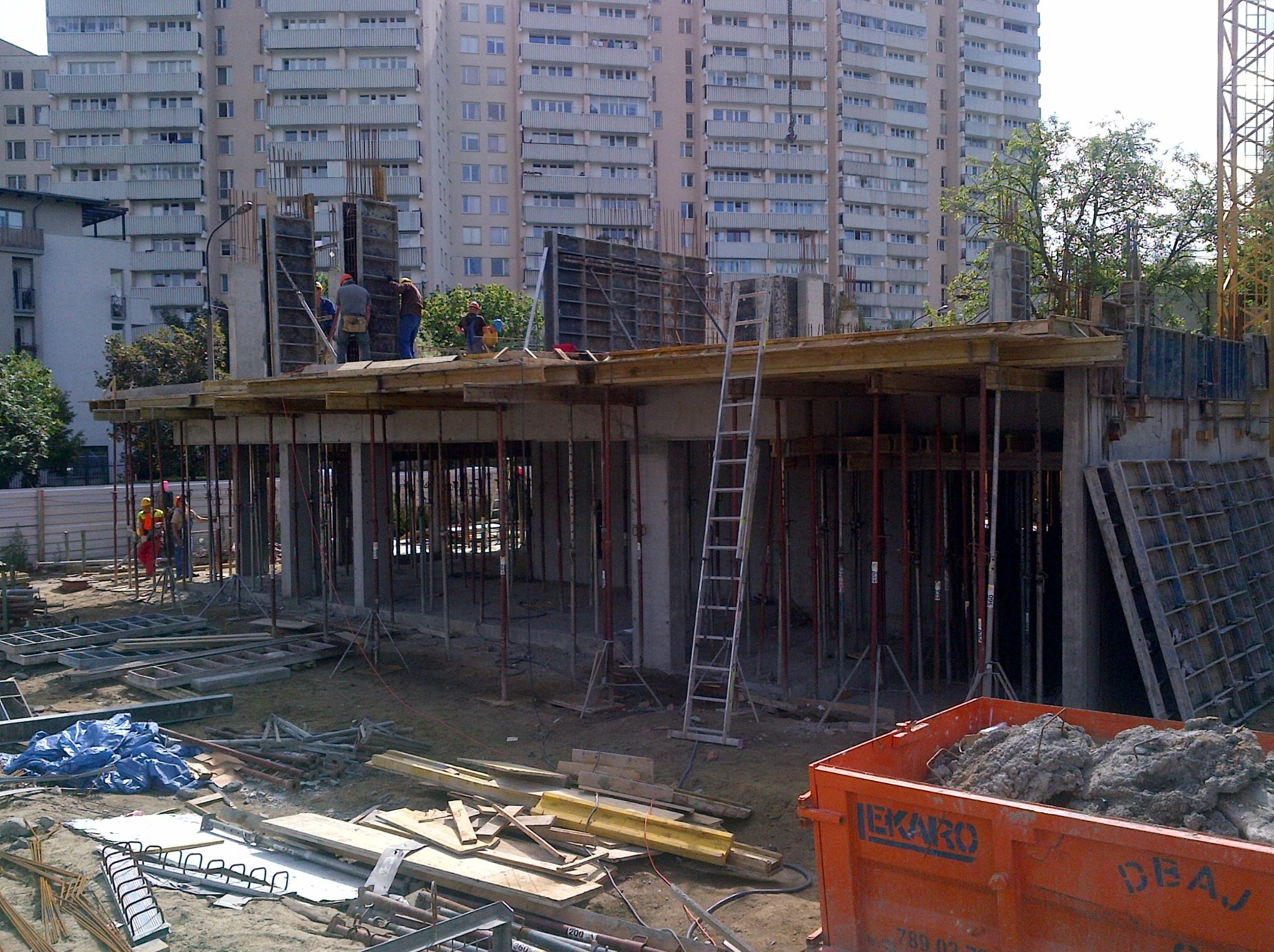 Budowa budynku żelbetowego w trakcie realizacji: widoczne szalunki, zbrojenia, podpory i pracownicy na tle wysokiego bloku mieszkalnego. Kontener na gruz w pierwszym planie.