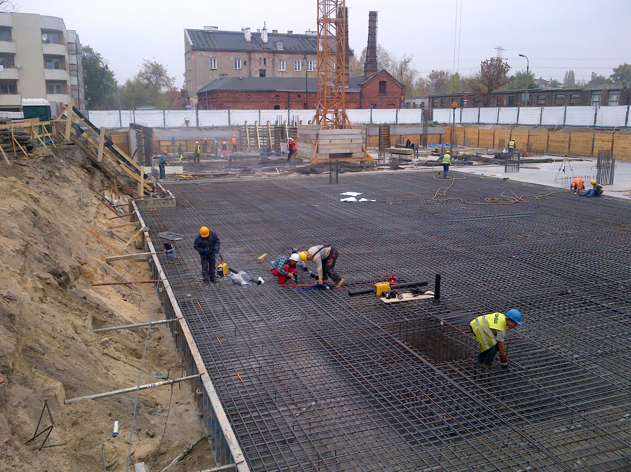 Zbrojenie fundamentów na budowie z widocznymi pracownikami w kaskach i kamizelkach odblaskowych. W tle budynki i żuraw budowlany.