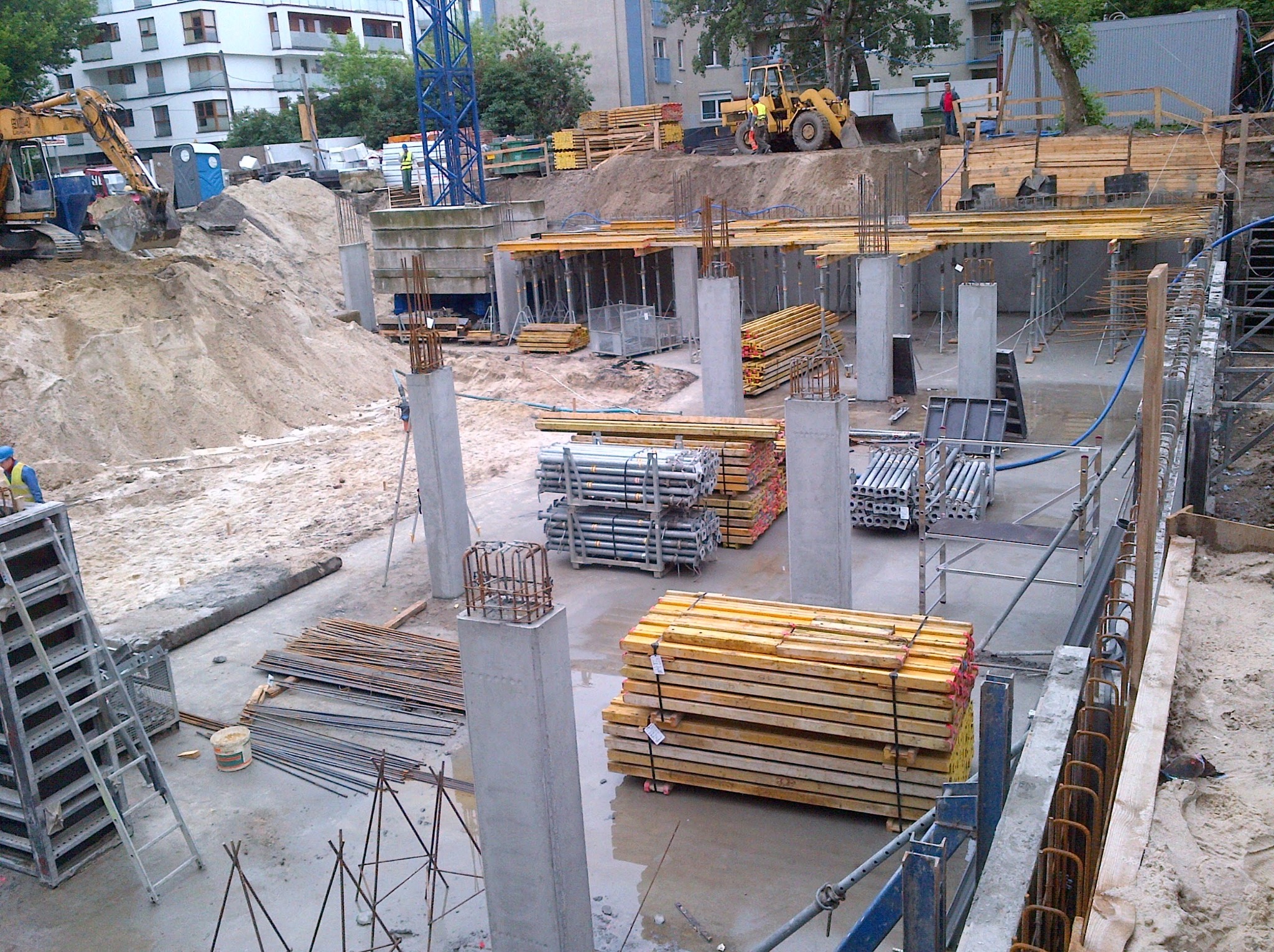 Wysoki kąt: plac budowy z betonowymi kolumnami, zbrojeniami, stertami drewna i sprzętem budowlanym w tle. Widoczne elementy szalunków i przygotowania do wylewania betonu.