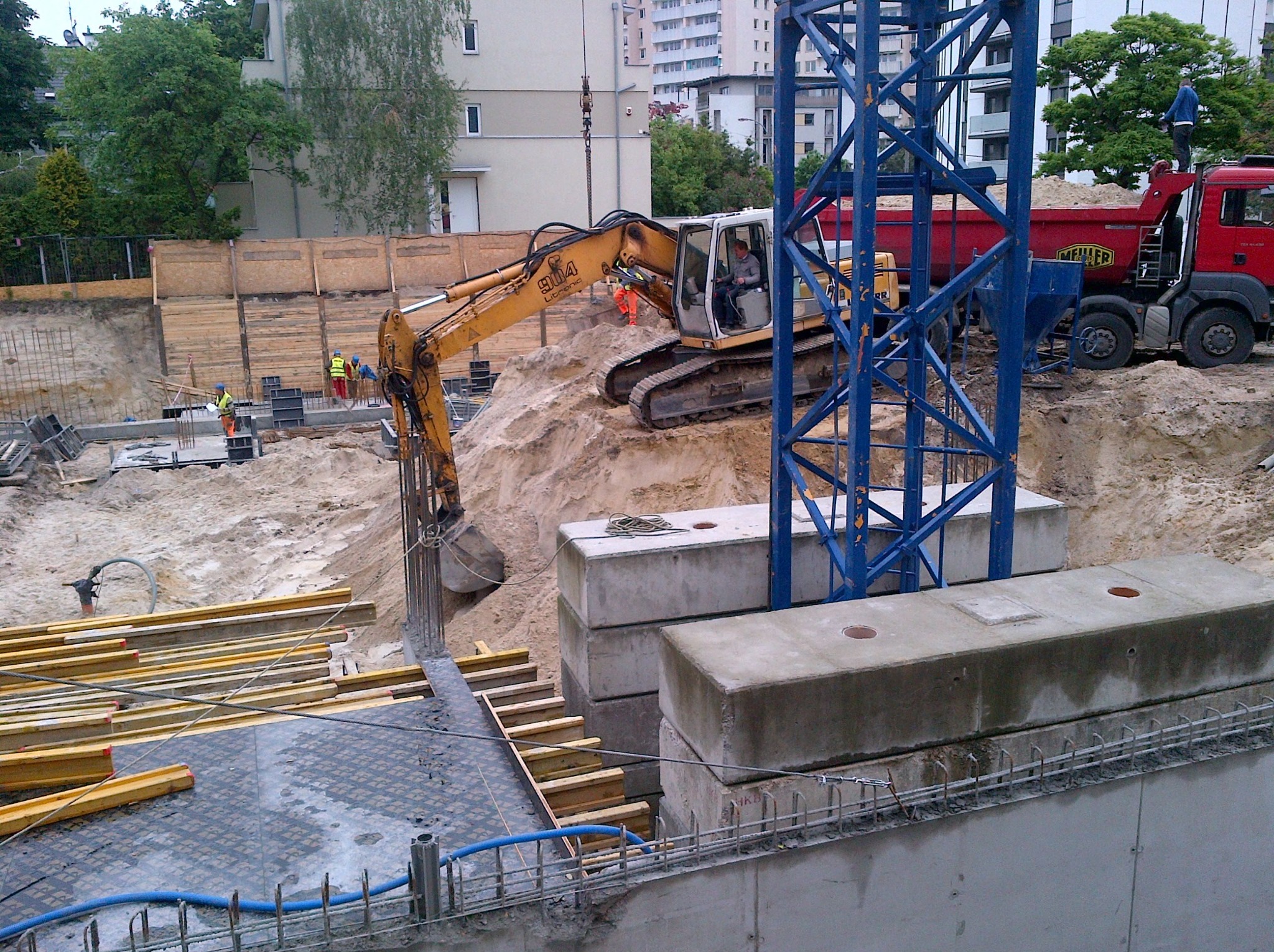 Żółta koparka na placu budowy, wbija łyżkę w ziemię obok betonowych bloków. W tle widoczni pracownicy i ciężarówka z piaskiem. Konstrukcja żurawia w kadrze.