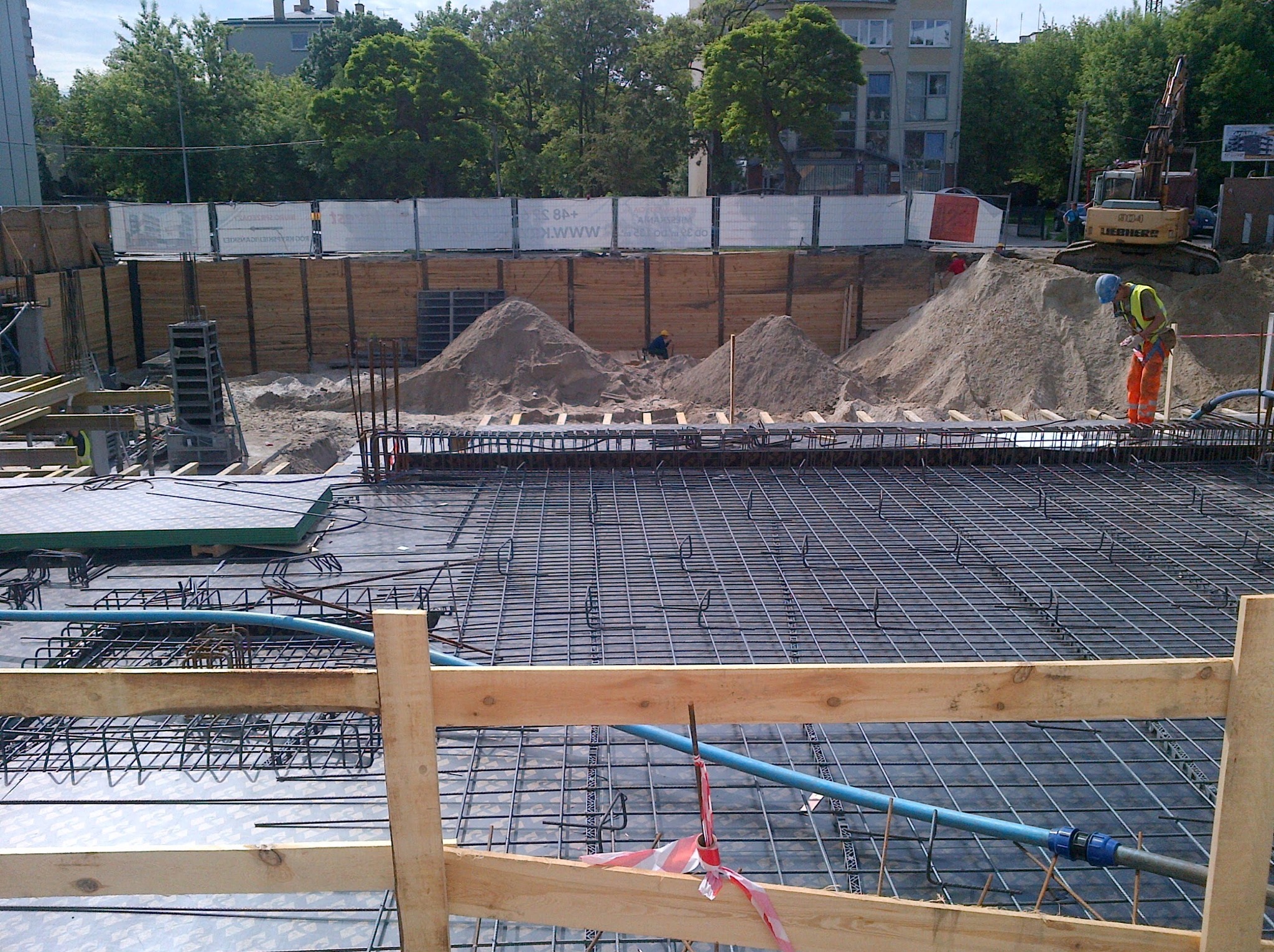 Siatka zbrojeniowa na budowie, widoczny pracownik w pomarańczowym kombinezonie i żółtej kamizelce, oraz koparka Liebherr w tle. Ujęcie z góry, w dzień.