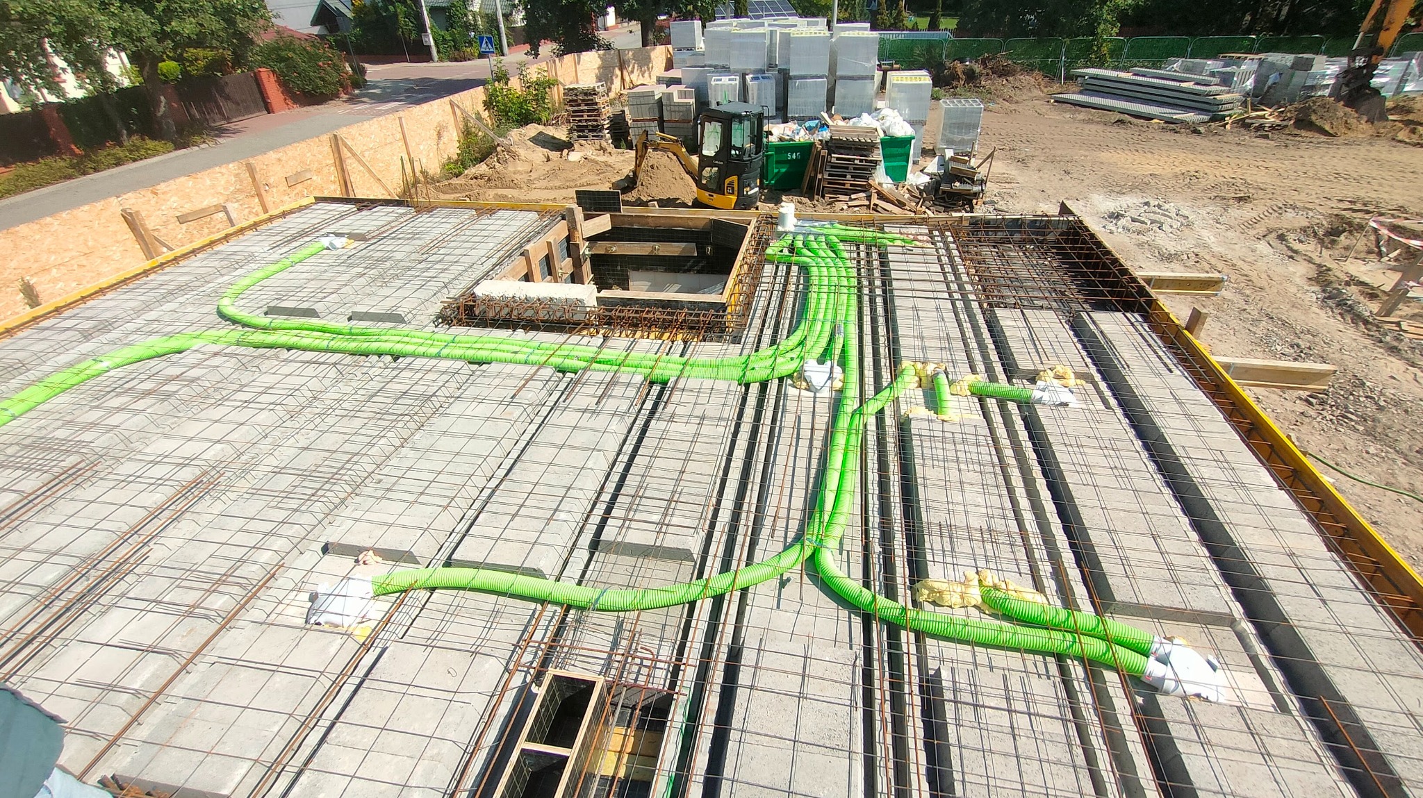 Zbrojenie stropu z zielonymi rurami instalacji elektrycznej, widok z góry na plac budowy z mini koparką i materiałami budowlanymi w tle, w słoneczny dzień.