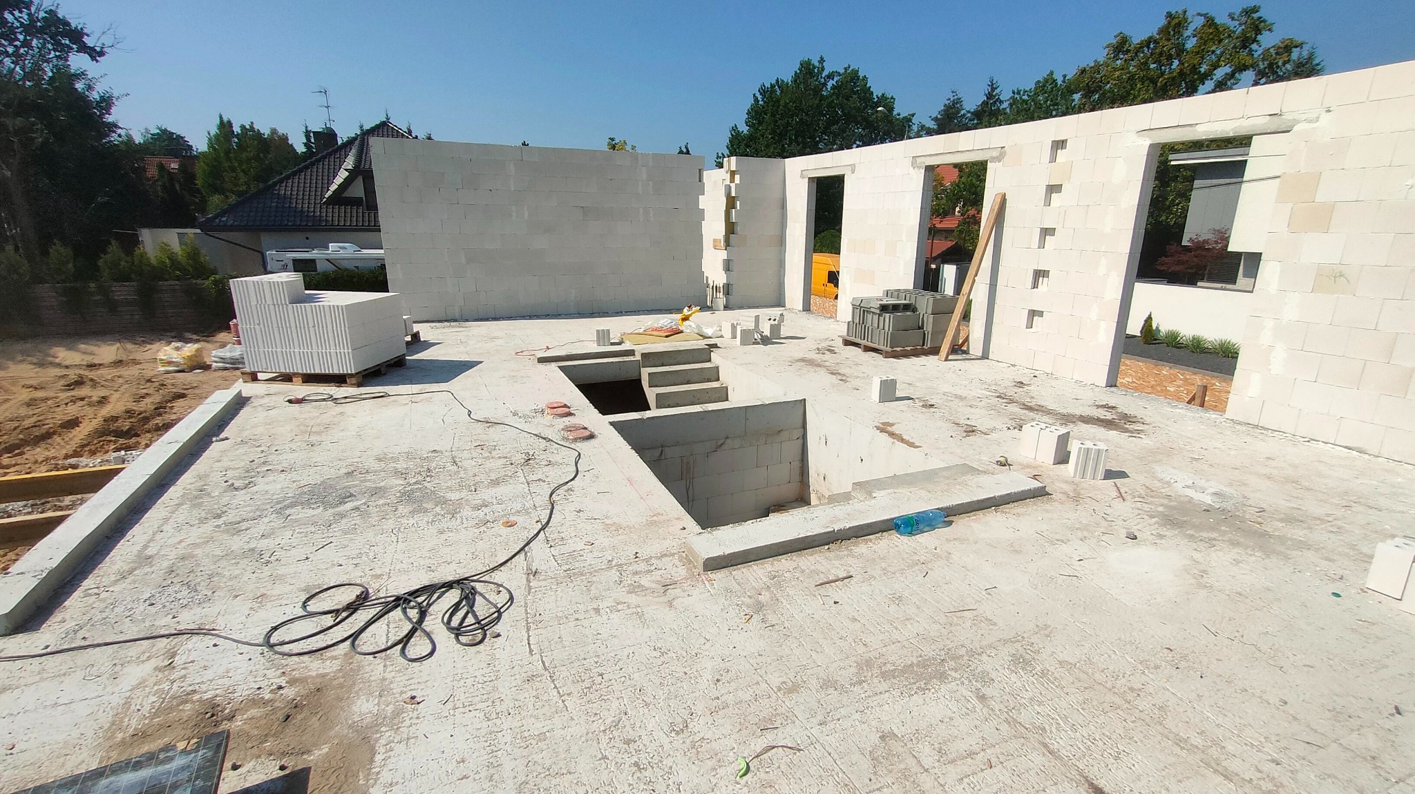 Wnętrze domu w trakcie budowy z betonu komórkowego. Widoczne ściany, otwory okienne i schody do piwnicy. Na podłodze leżą materiały budowlane i narzędzia.