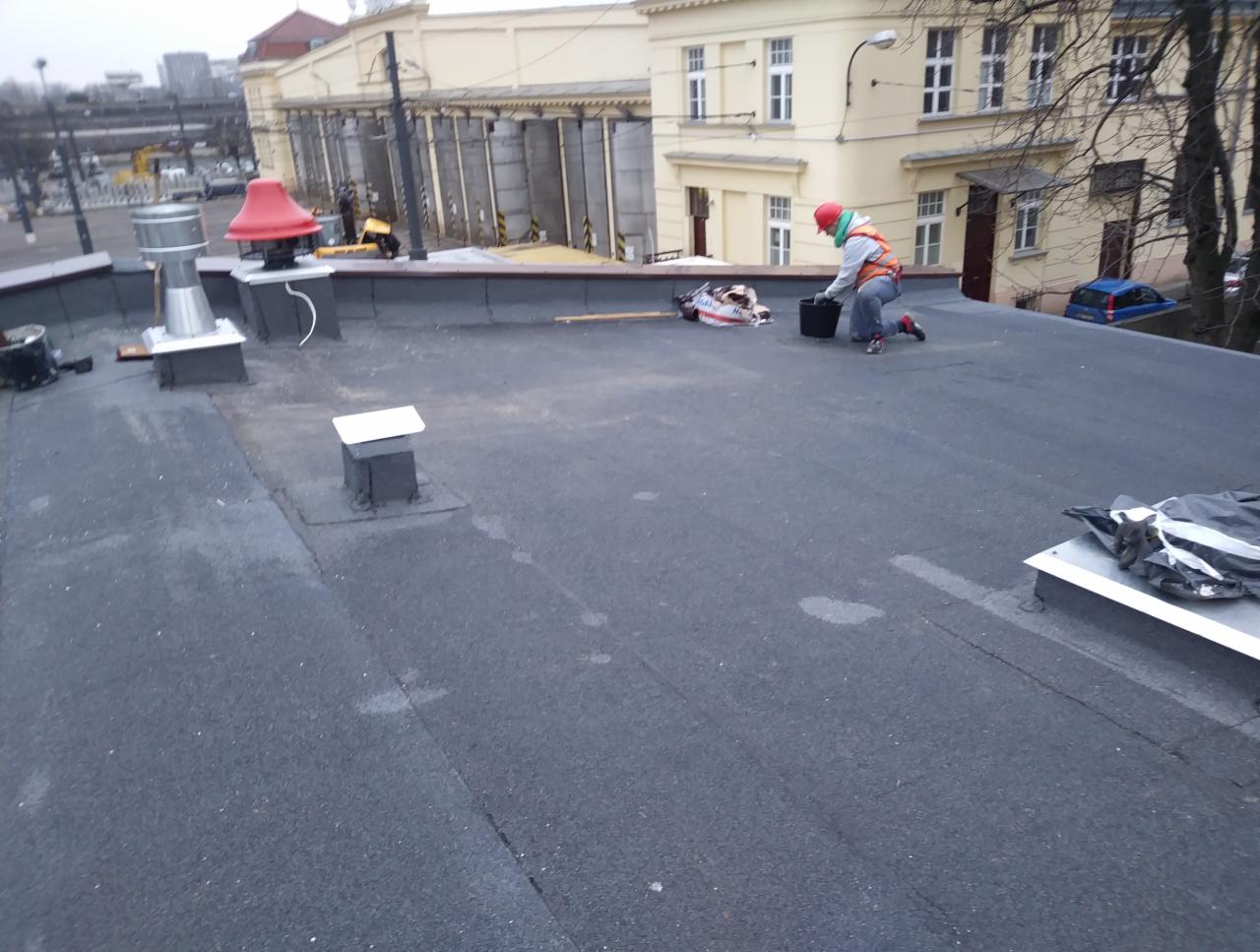 Pracownik w kasku i kamizelce odblaskowej pracuje na płaskim dachu pokrytym papą, w tle budynek z kolumnami i fragment miasta.