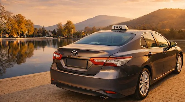 Szary samochód taxi Toyota Camry z lampą TAXI na dachu, zaparkowany nad brzegiem jeziora o zachodzie słońca. W tle góry i jesienne drzewa.