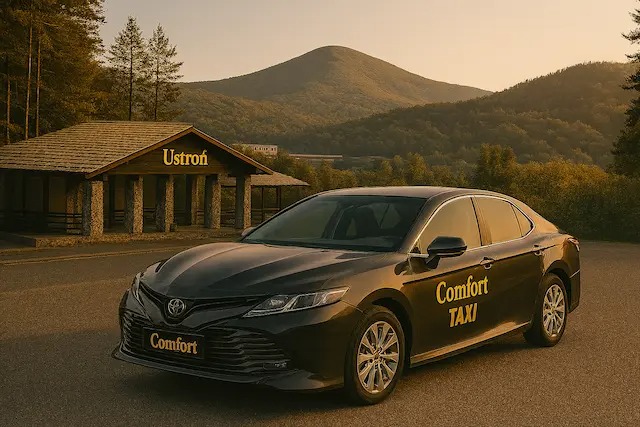 Ciemny sedan Toyota Camry z napisem 'Comfort TAXI' na tle budynku z napisem 'Ustroń' i gór w tle, w ciepłym świetle zachodzącego słońca.