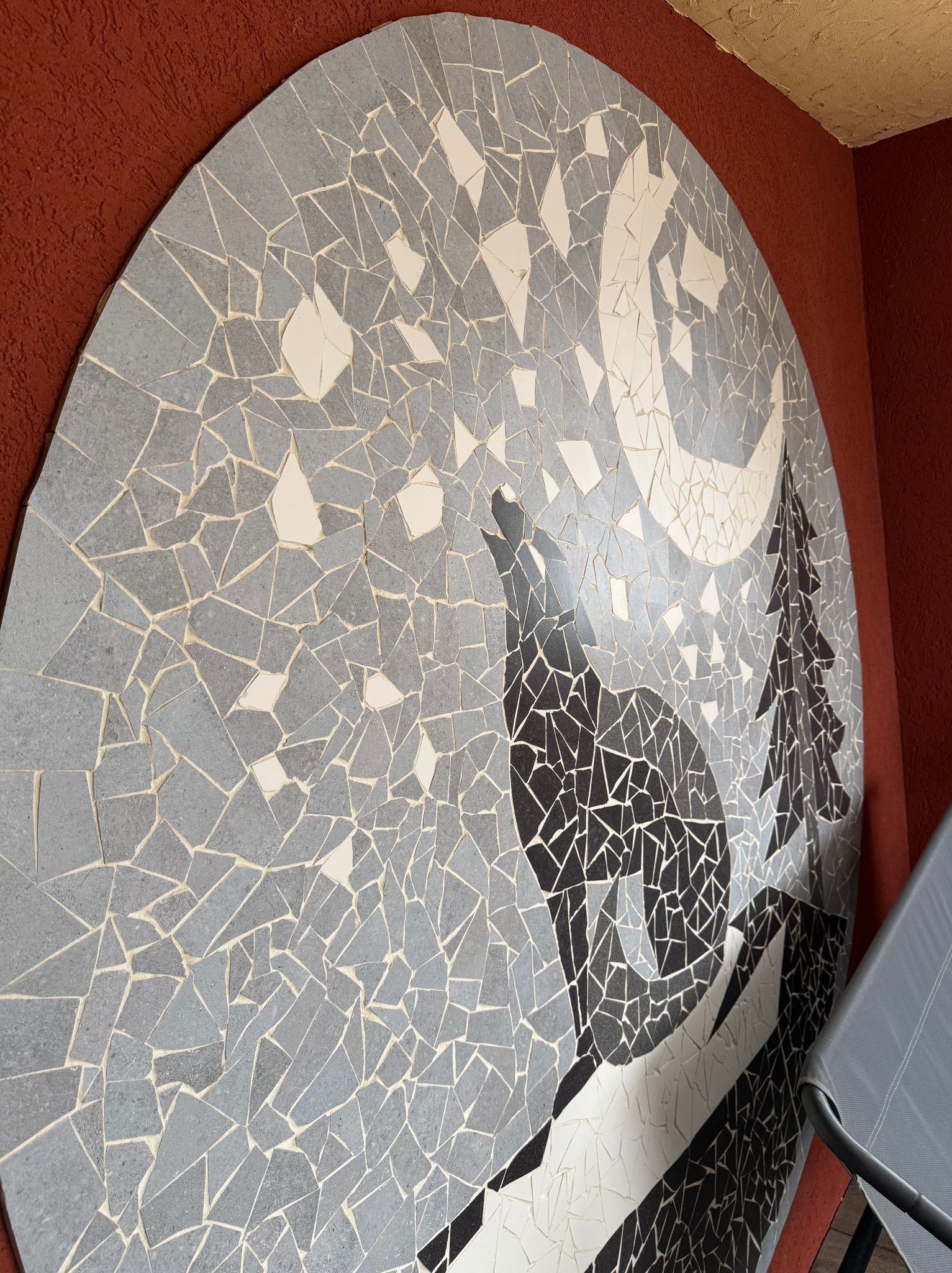 Mozaika ścienna z motywem wilka wyjącego do księżyca i drzewa iglastego, wykonana z szarych, białych i czarnych płytek na czerwonej ścianie. Widoczny fragment krzesła.