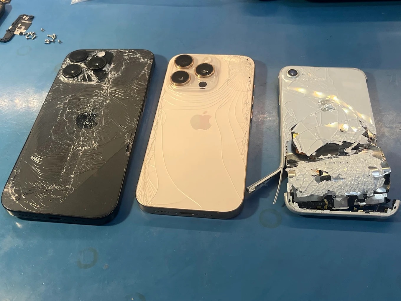 Trzy uszkodzone telefony iPhone z potłuczonymi ekranami i obudowami, leżące obok siebie na niebieskim blacie w serwisie we Wrocławiu. Widoczne drobne elementy demontażu.
