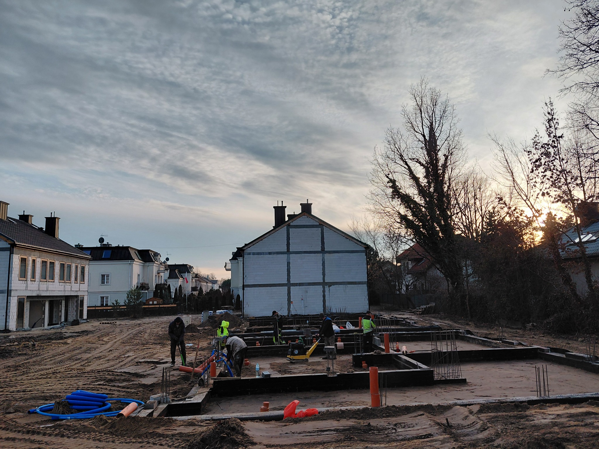 Budowa fundamentów domu jednorodzinnego w trakcie prac. Widoczni pracownicy w odblaskowych kamizelkach i sprzęt budowlany. W tle budynek w stanie surowym otwartym.