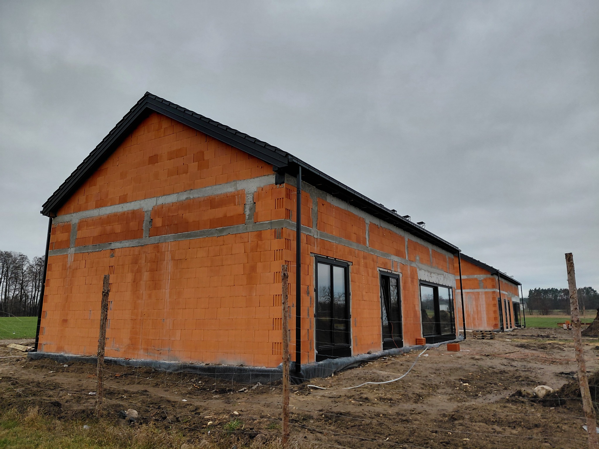 Budowa domu z czerwonej cegły ceramicznej, widoczne okna z czarnymi ramami i fragment dachu. Teren budowy ogrodzony, pochmurne niebo w tle.