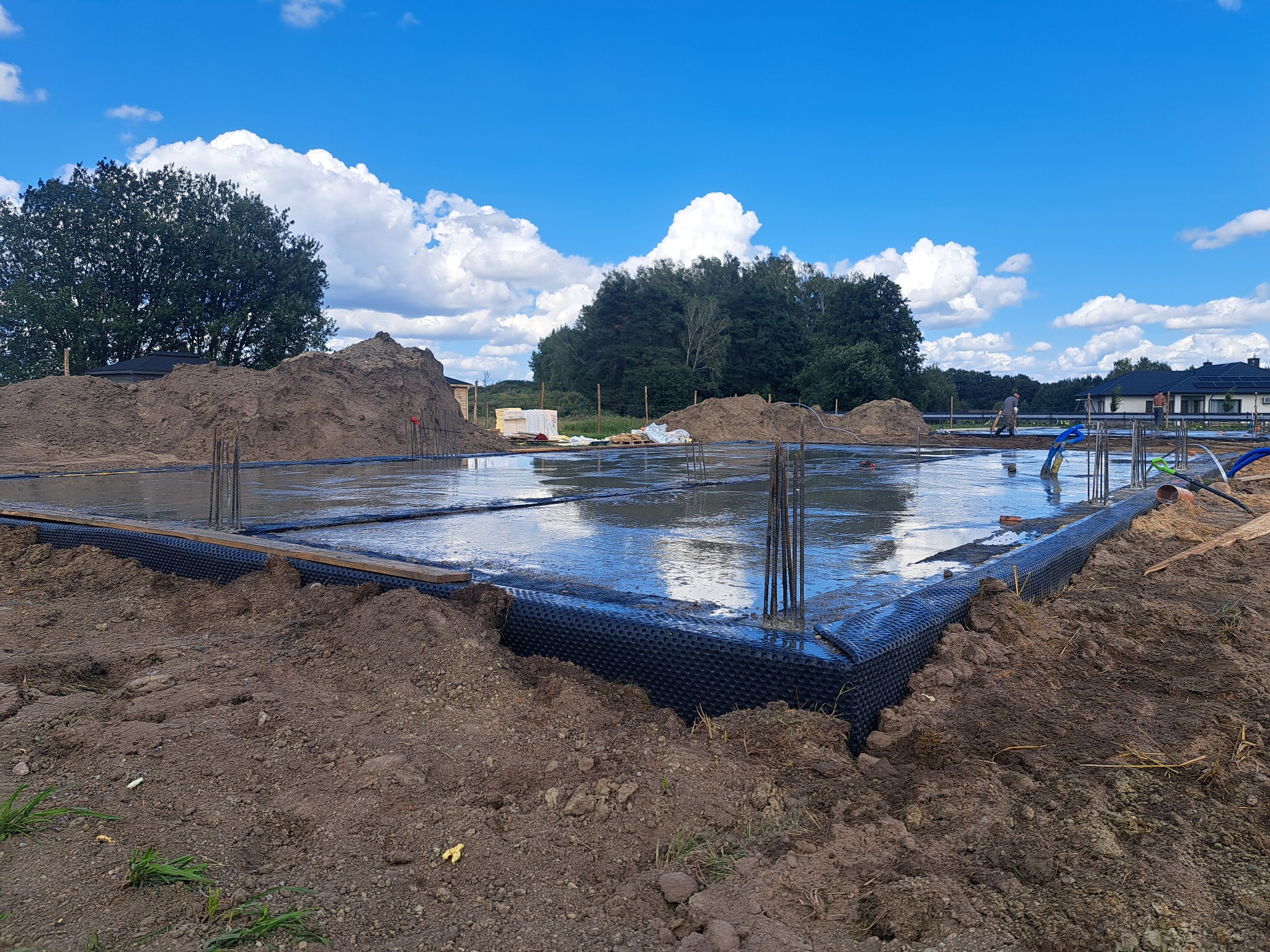 Zalane fundamenty domu jednorodzinnego z wystającymi prętami zbrojeniowymi, zabezpieczone folią kubełkową, na tle błękitnego nieba i zieleni.