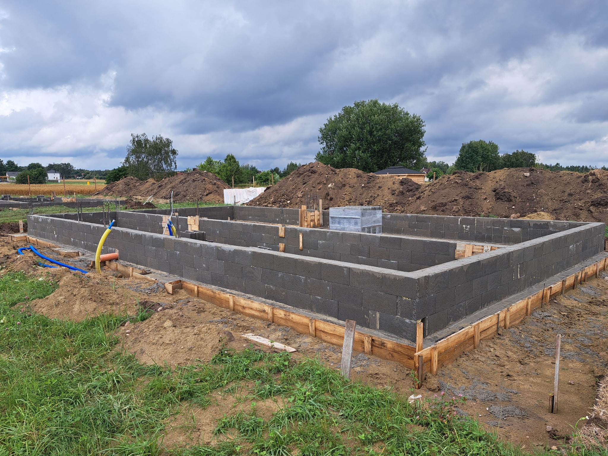 Fundamenty domu z bloczków betonowych, szalunek drewniany, widoczne instalacje, ziemia, drzewa w tle, pochmurne niebo. Etap budowy.