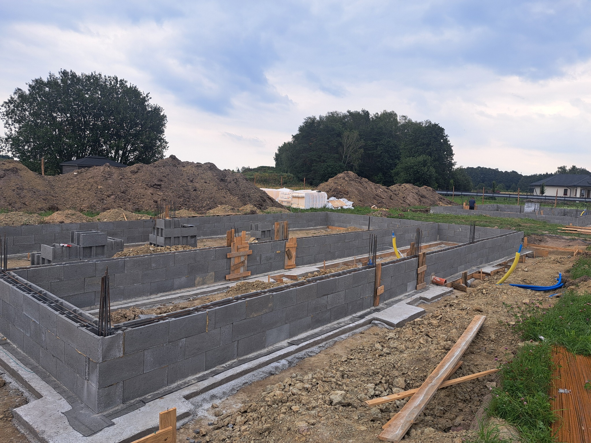 Fundamenty domu z bloczków betonowych, zbrojenie pionowe i poziome, szalunki drewniane, teren budowy z hałdami ziemi i zielenią w tle, pochmurne niebo.