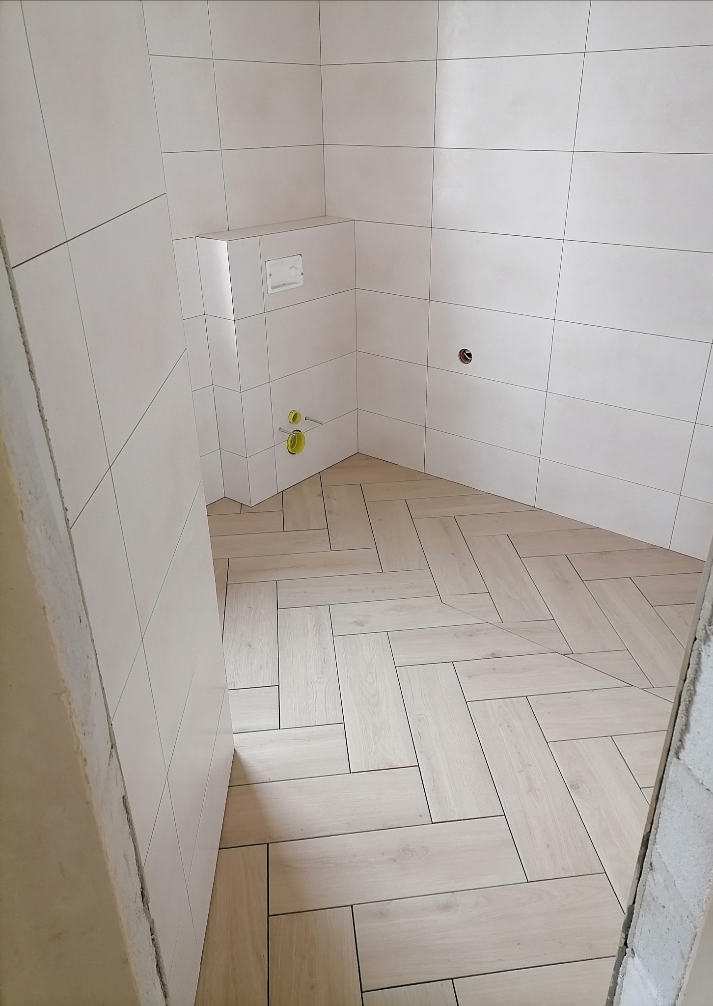 Jasna łazienka po remoncie: białe płytki ścienne i podłoga w jodełkę z paneli imitujących drewno. Widoczne przyłącza wodne i fragment surowego wejścia.