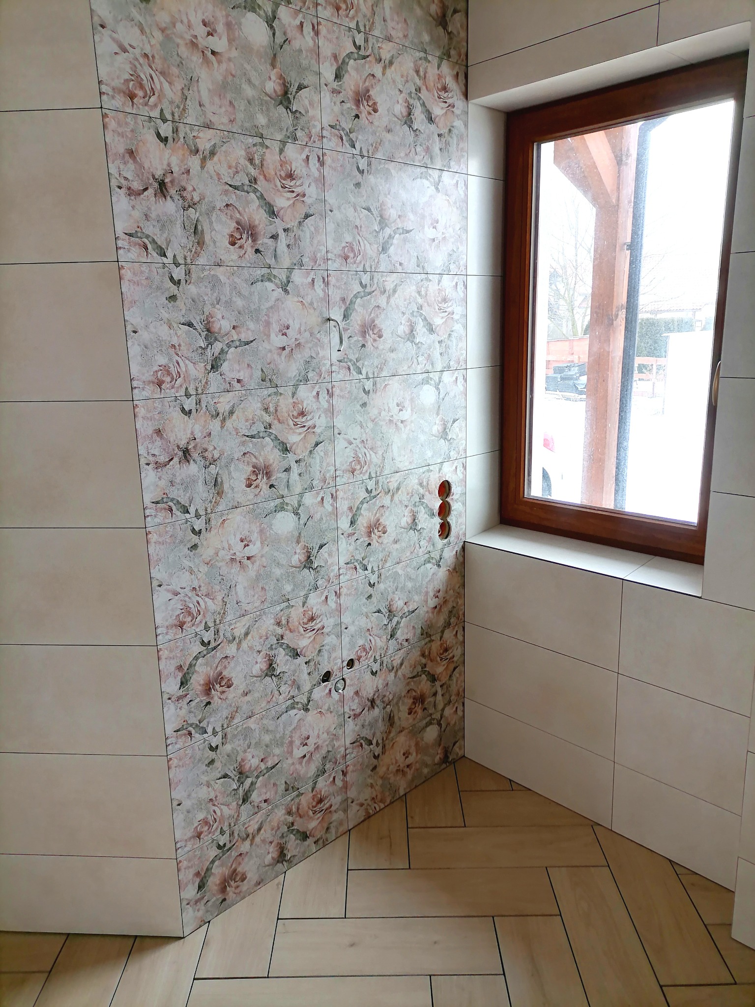 Ukończona łazienka z kwiatowym wzorem na ścianie, jasnymi płytkami i drewnianą ramą okienną. Widoczne gniazdka elektryczne i ułożona podłoga w jodełkę.