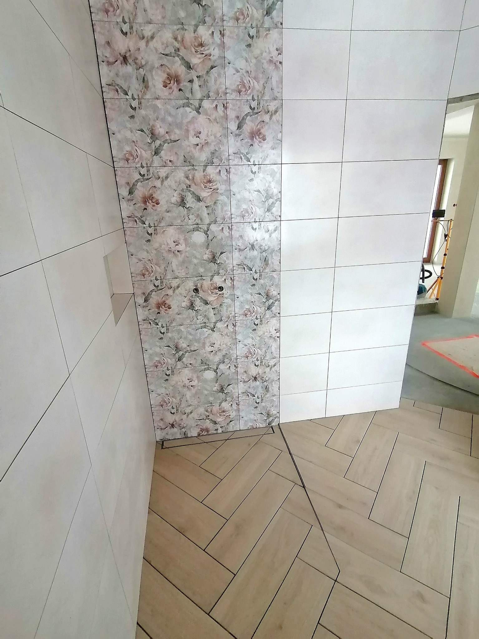 Ukończona ściana z płytek z motywem kwiatowym, kontrastująca z białymi płytkami i podłogą w jodełkę. Widoczny fragment pomieszczenia w trakcie wykańczania.