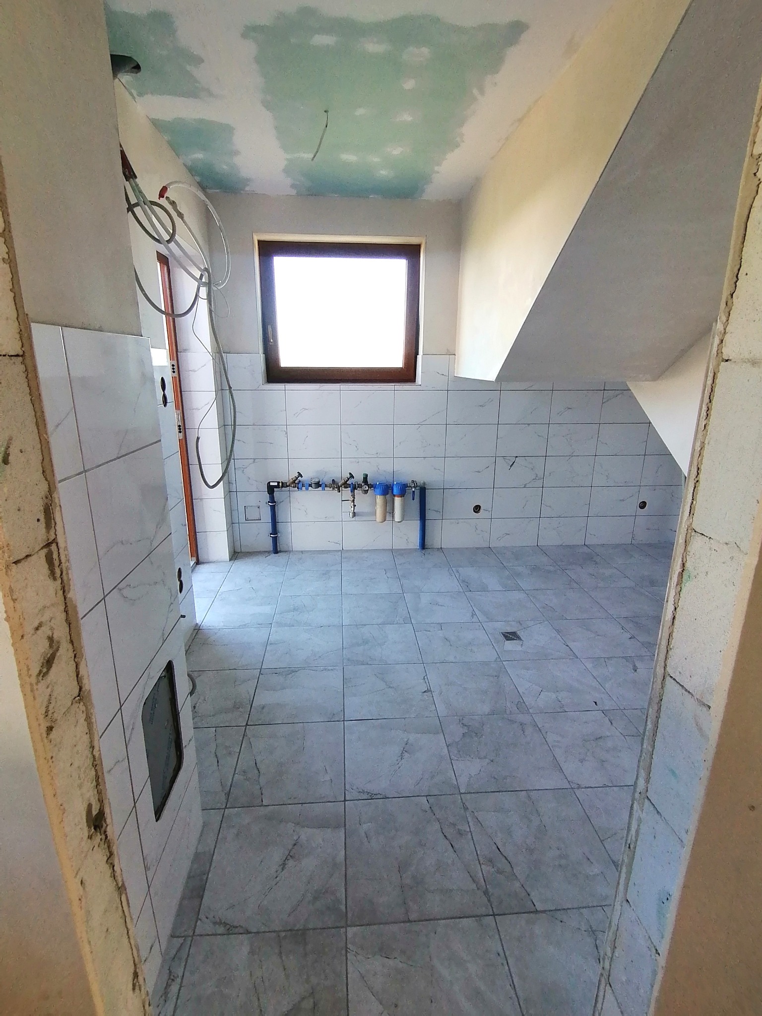 Wnętrze łazienki w trakcie prac wykończeniowych: ułożone płytki podłogowe i ścienne, widoczne instalacje wodne, sufit z widocznymi śladami szpachlowania.