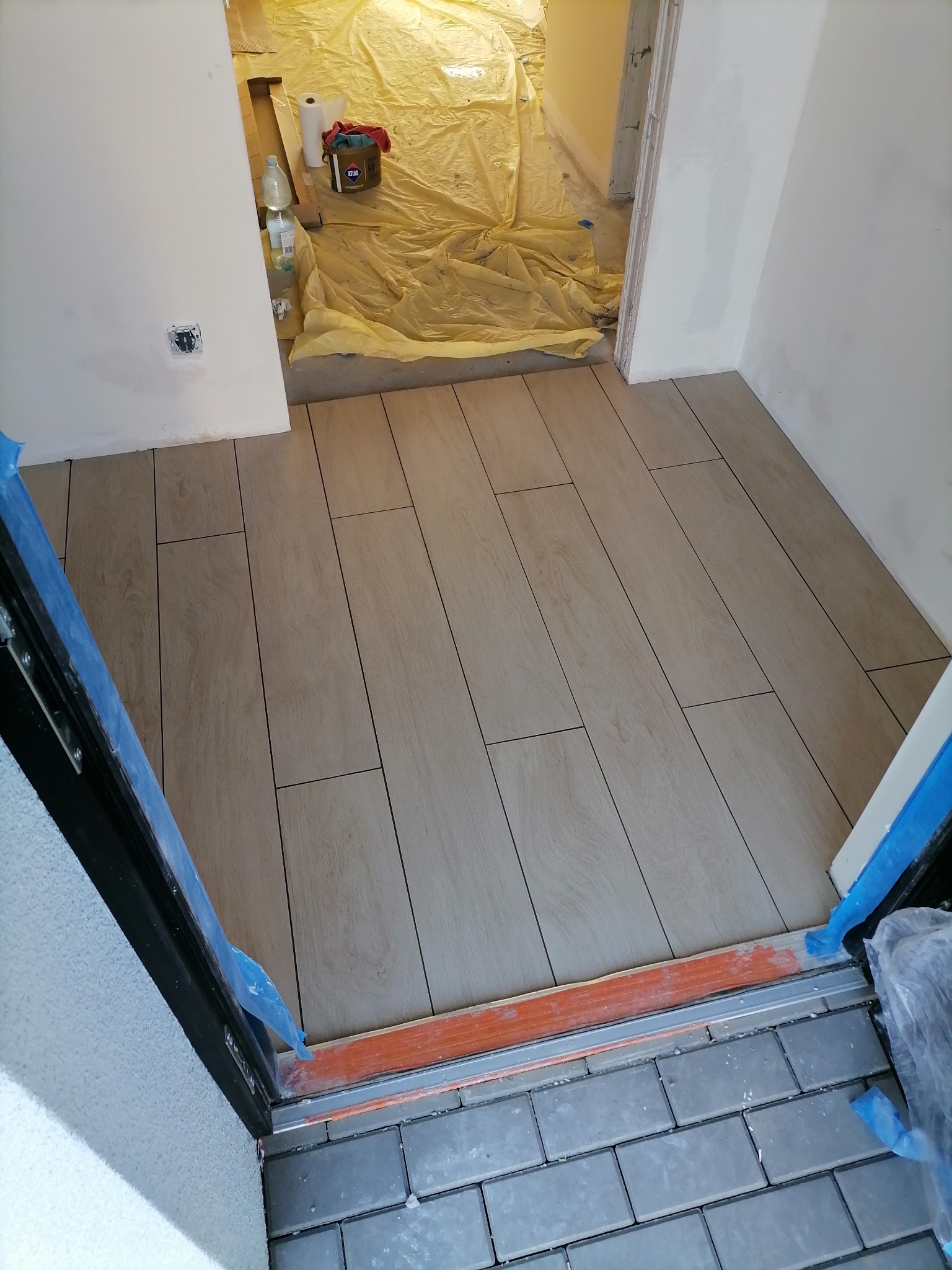 Ułożona podłoga z płytek imitujących drewno w korytarzu, widok z góry, z zabezpieczonym wejściem i widocznym pomieszczeniem w trakcie prac wykończeniowych.