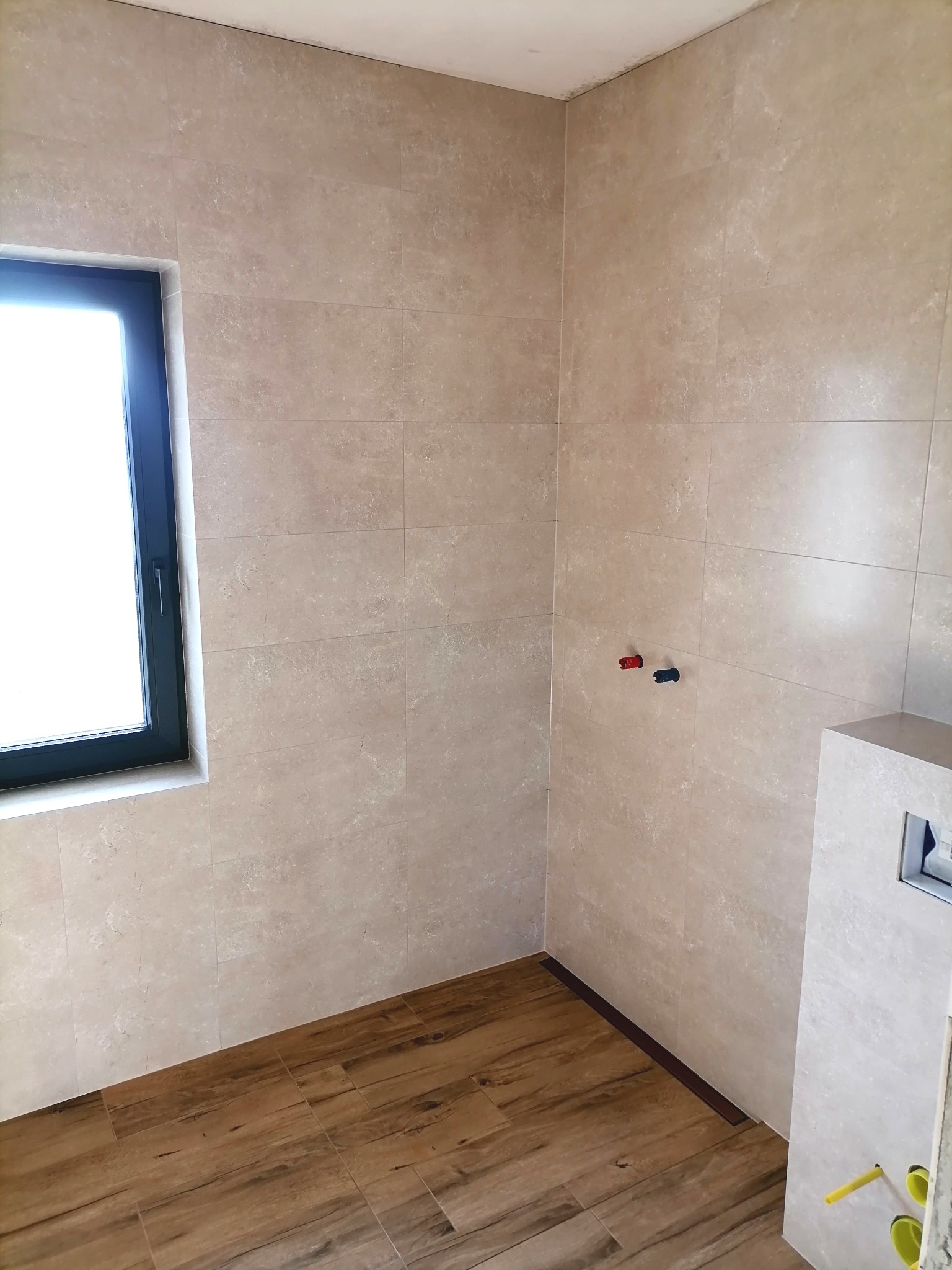 Jasne, beżowe płytki ścienne w łazience z oknem i drewnopodobną podłogą. Widoczne przyłącza wodne i fragment zabudowy WC.