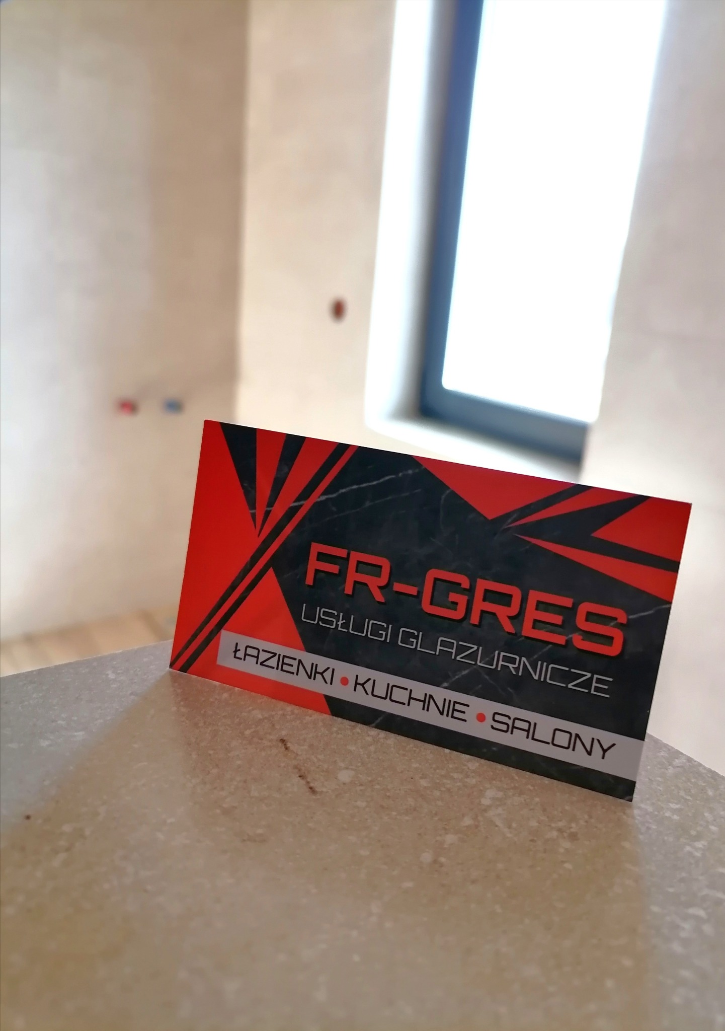 Wizytówka firmy FR-GRES na tle wykończonej łazienki z oknem dachowym. Usługi glazurnicze: łazienki, kuchnie, salony. Czerwono-czarna grafika.