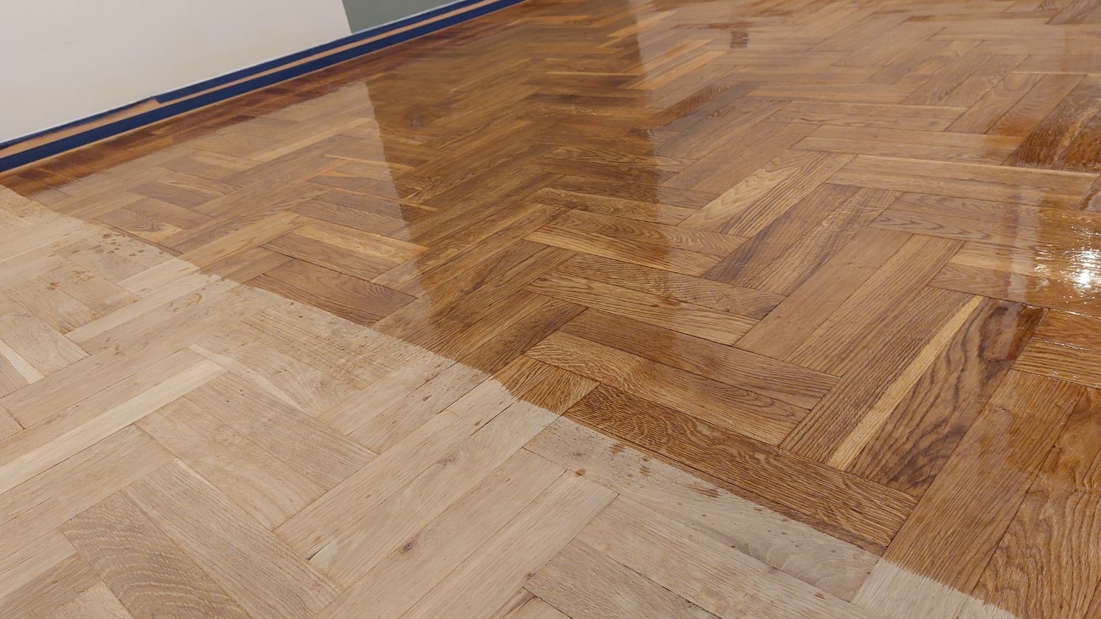 Parkiet w jodełkę, częściowo odnowiony, z widoczną różnicą w kolorze i połysku między starą a nową powierzchnią. Świeżo nałożony lakier podkreśla strukturę drewna.